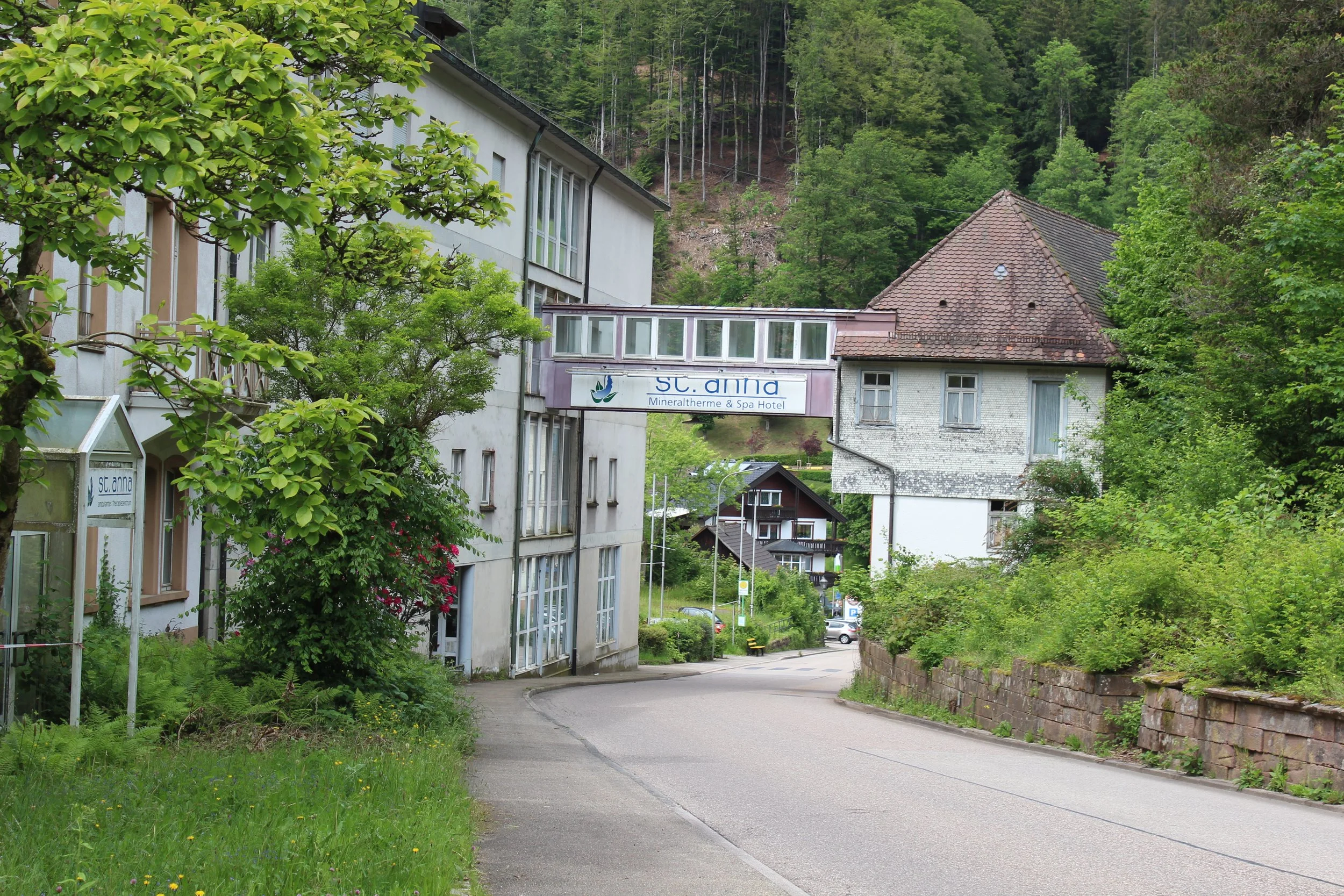 Eine Straße in einer grünen, bergigen Gegend mit Gebäuden, darunter ein Spa-Hotel namens 'St. Anna' und mehrere kleine Häuser, umgeben von Bäumen und Hügeln.