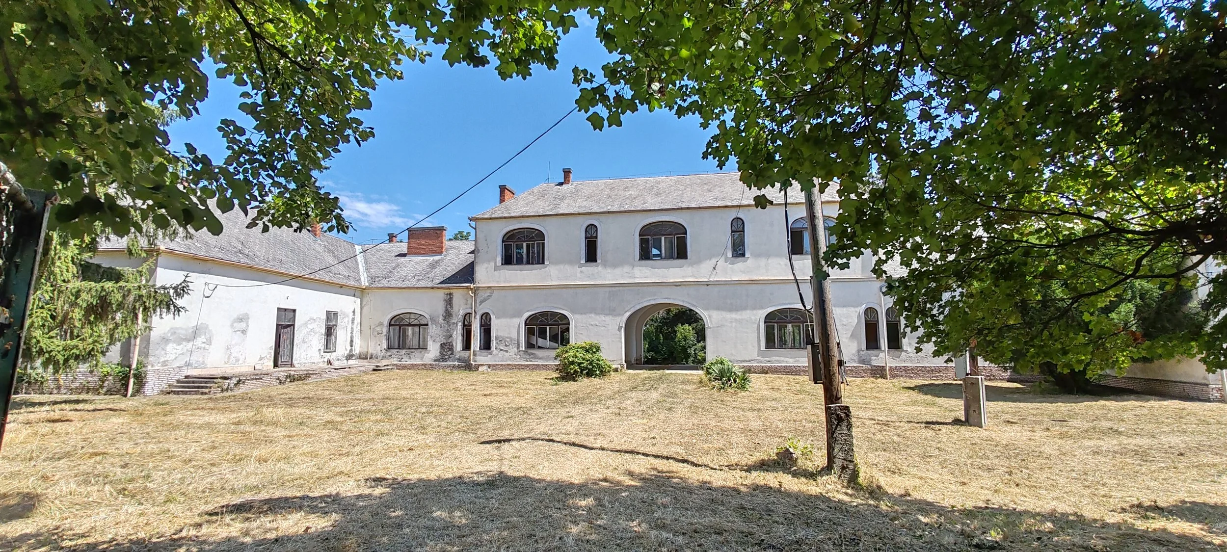 Ein altes, weißes Gebäude mit mehreren Fenstern und einer Arkade, umgeben von Bäumen und einem Rasen.