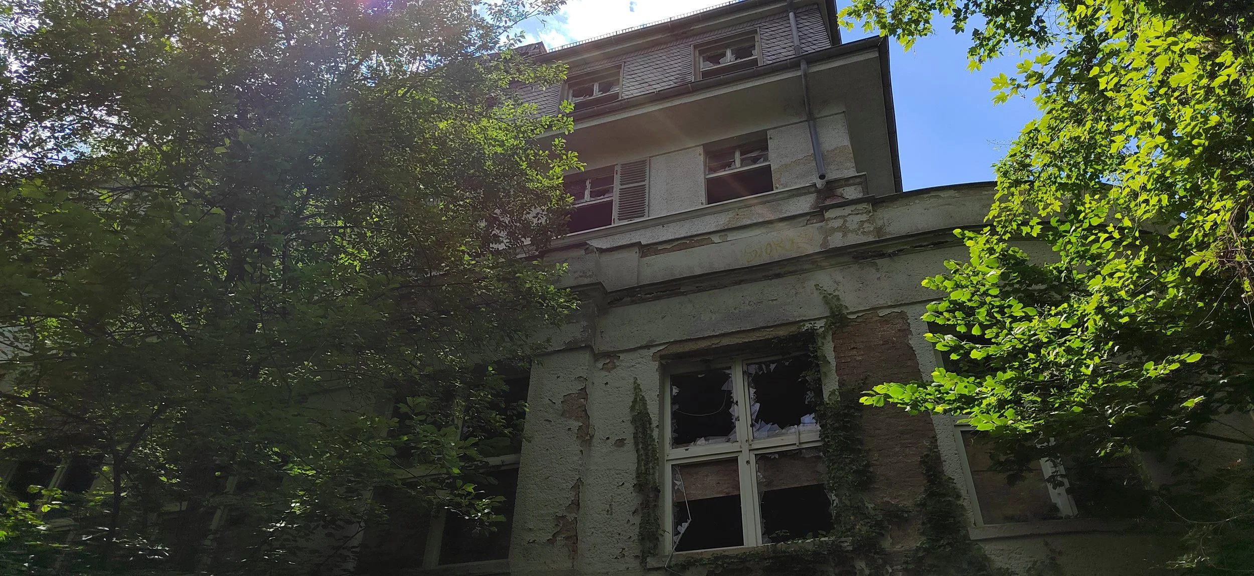 Verfallenes Gebäude, teilweise von Bäumen umgeben, mit beschädigten Fenstern und sichtbaren Abrieb an den Wänden, bei sonnigem Himmel.