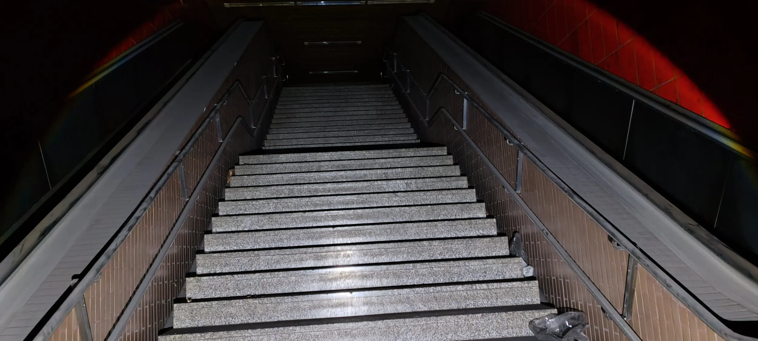 Treppennach unten in einer unterirdischen Station, mit grauen Stufen,br/  Holzseitengeländer, rote Ziegelwände und dunkle Decke, leicht schmutzig und Müll am Boden.