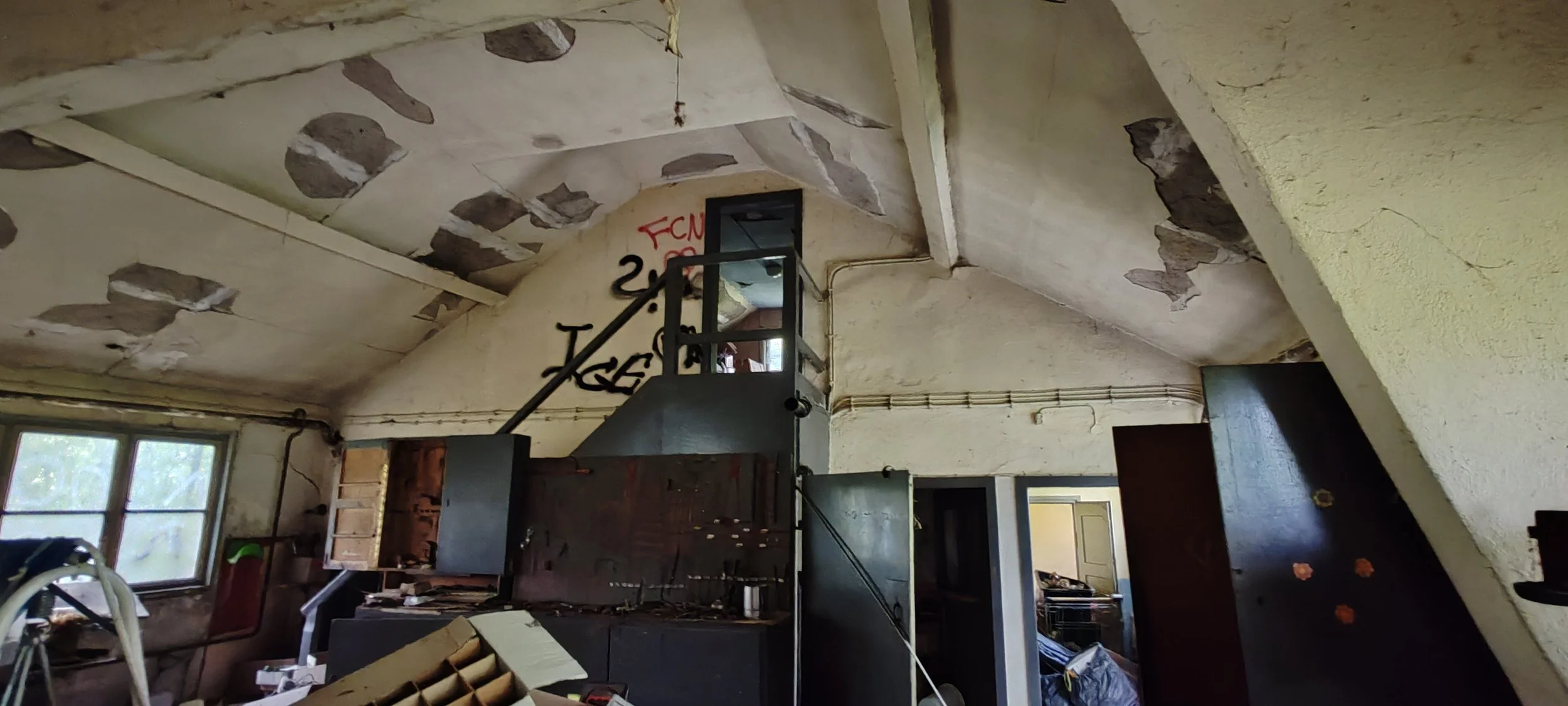 Ansicht eines alten, verfallenen Raumes mit schmutzigen, beschädigten Wänden und einer schmutzigen Fensterfront. Im Raum befinden sich alte Möbelstücke und Abfälle.