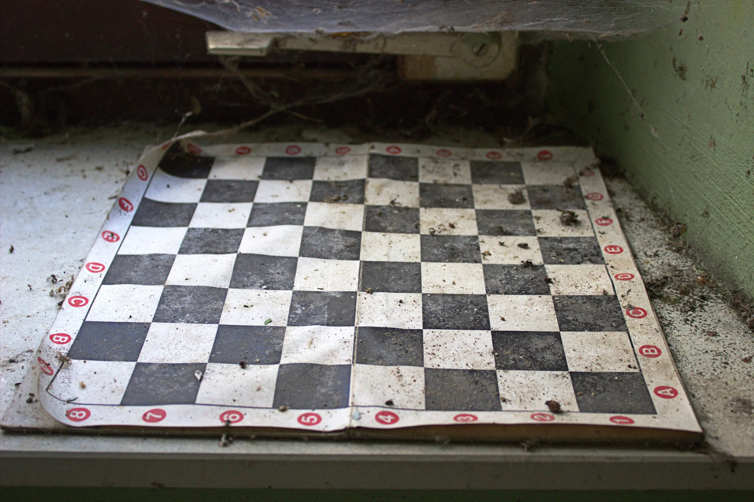 Ein verstaubtes, schmutziges Schachbrett liegt auf einer Fensterbank, umgeben von Staub und Spinnweben.