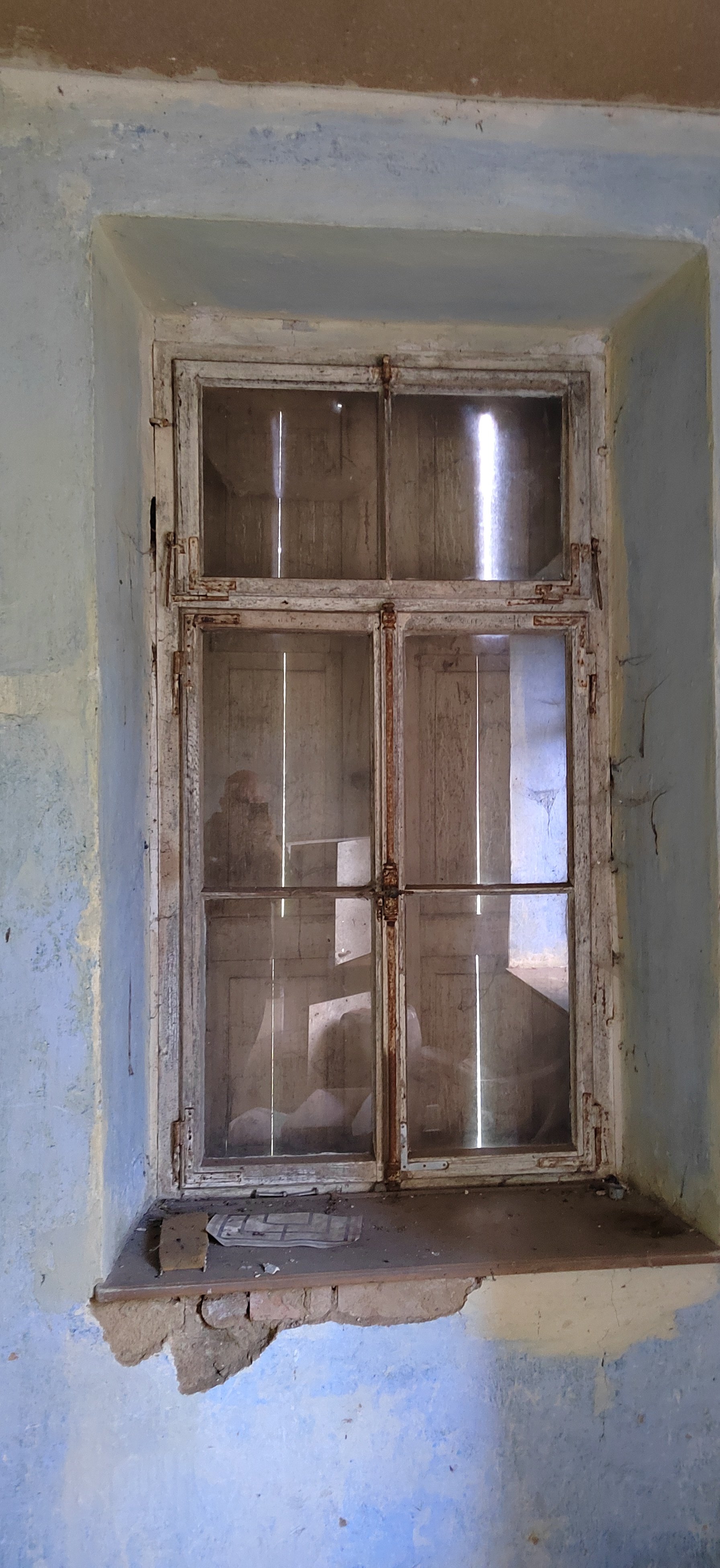 Ein alter, verrosteter Fensterrahmen mit sechs Scheiben in einer alten, abgenutzten Wand mit blauer und grüner Farbe.