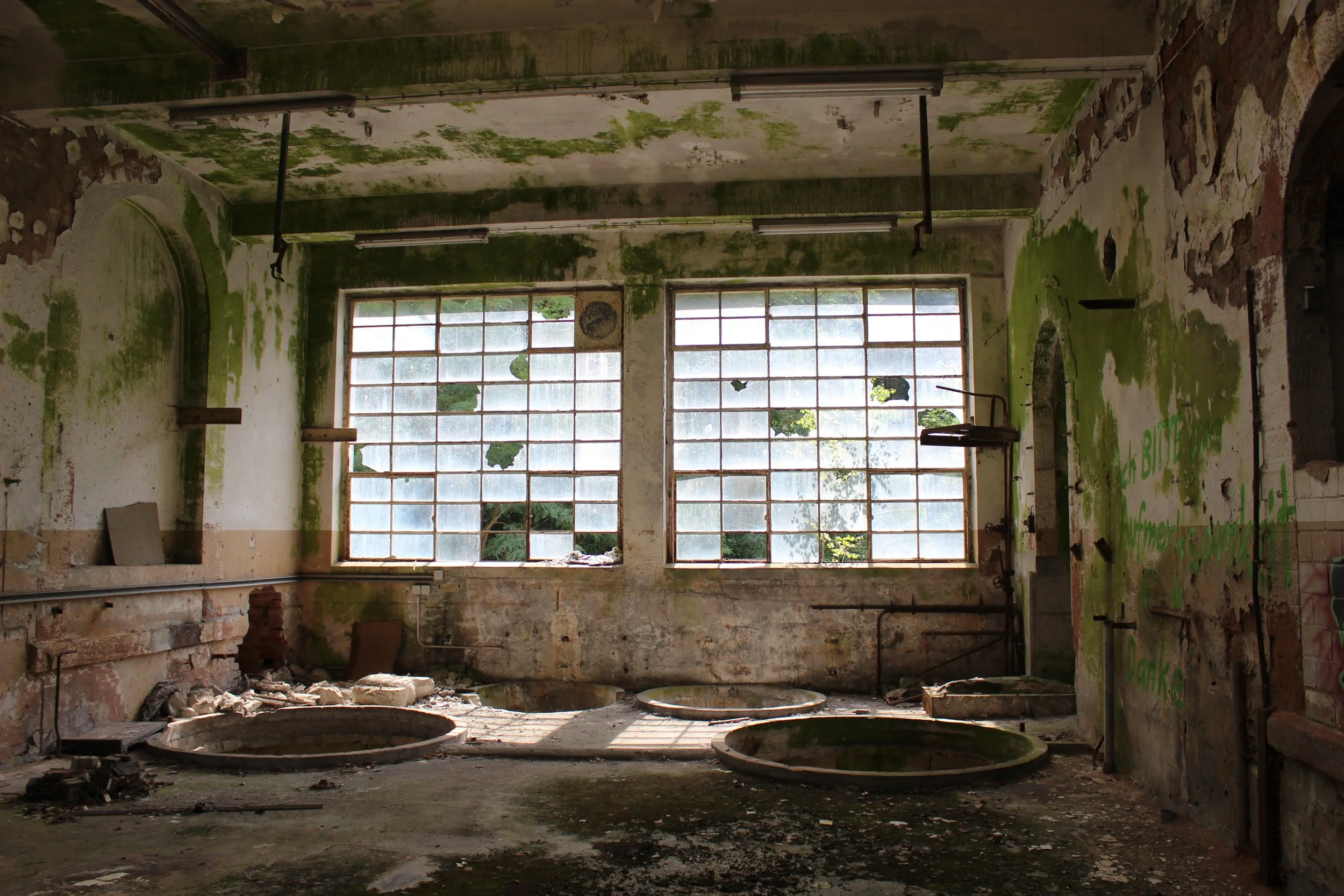 Ein verlassener, verfallener Raum mit großen schmutzigen Fenstern, grüner Moos- und Schmutzverschmutzung auf den Wänden, drei große, alte, bereits verrostete Waschbecken auf dem Boden, an den Wänden Graffiti in Grün, zerbrochene Wände, Staub und Schm