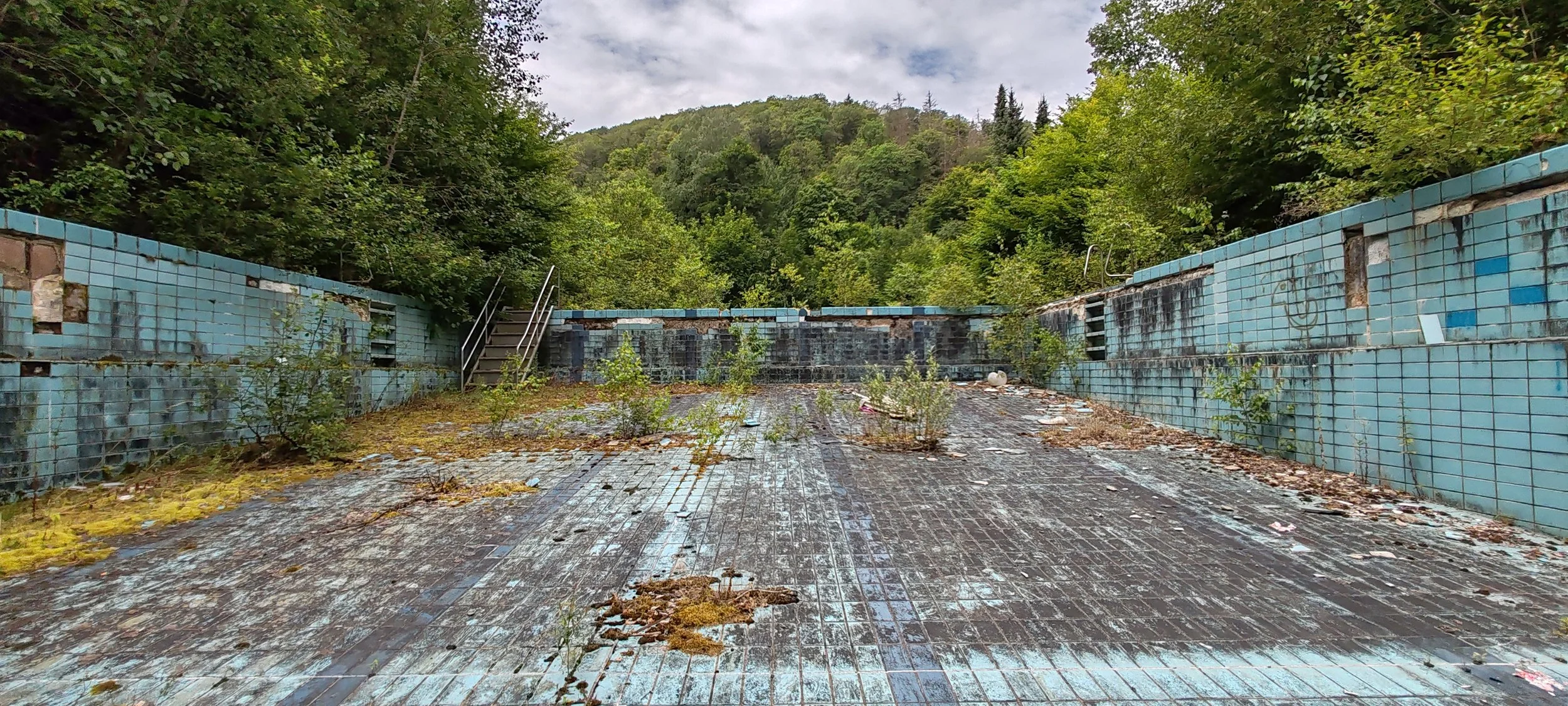 Verlassene, verfallene Schwimmbad mit überwucherten Fliesen und Pflanzen, im Hintergrund Bäume und bewölkter Himmel.