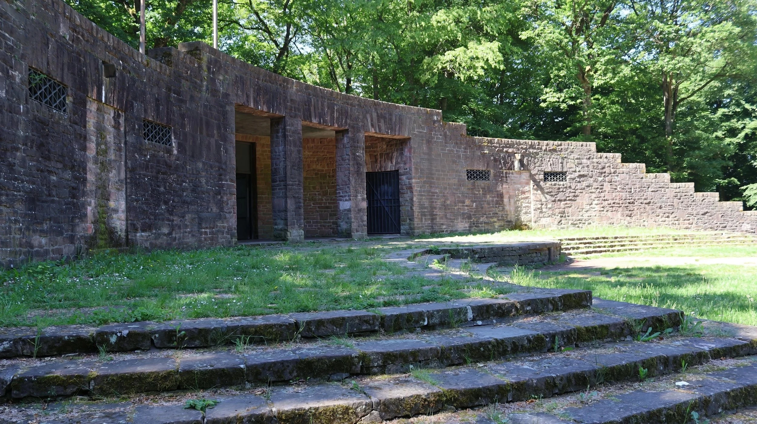 Steinerne Treppen vor einer Steinmauer auf einer Wiese, umgeben von grünen Bäumen.
