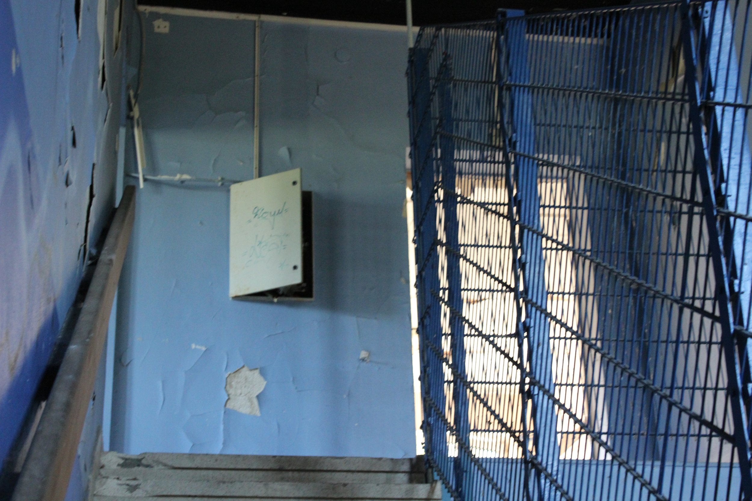 Veralteter, verlassen wirkender Treppenabsatz mit blauer Wand, zerrissener Tapete und einem geöffneten, weißen Briefkasten, der sich an der Wand befindet.