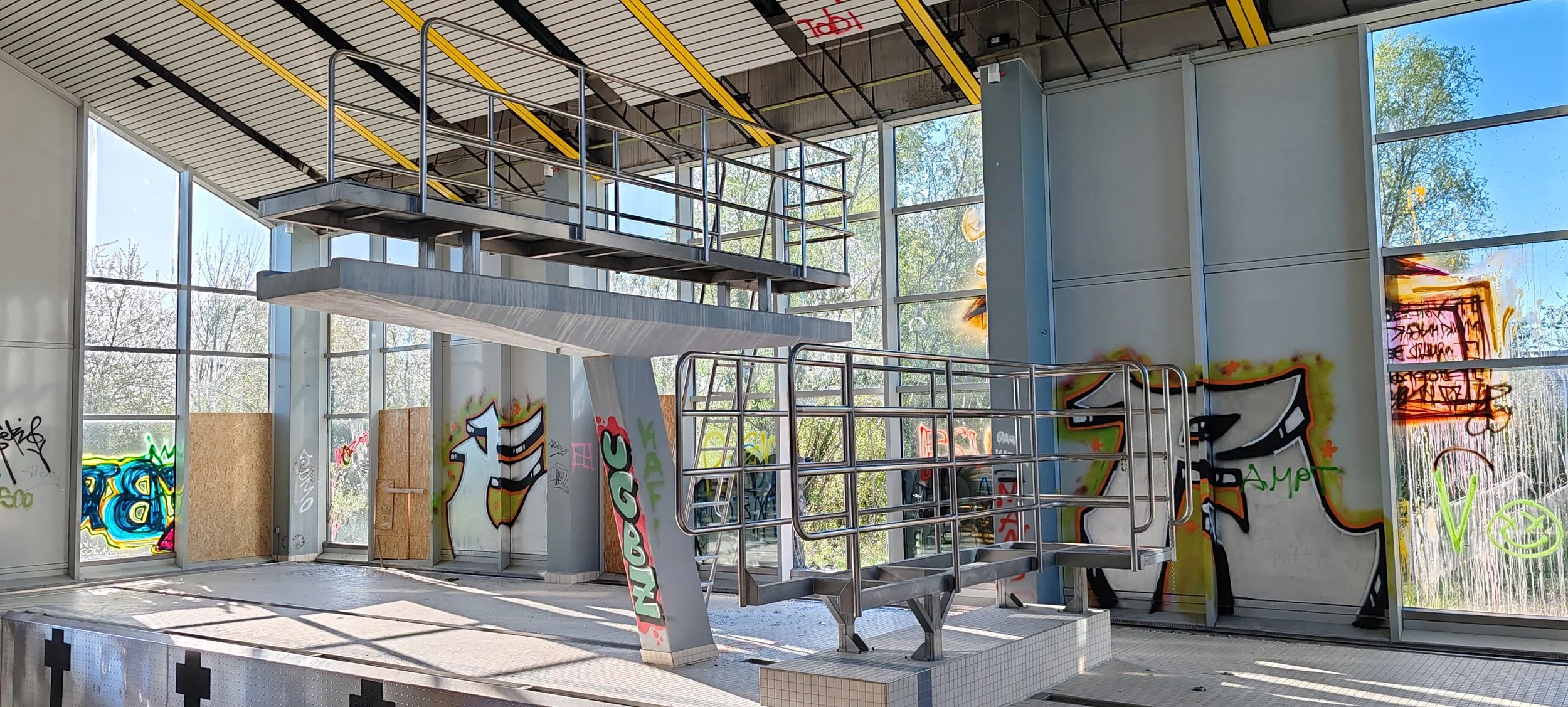 Leerer Swimmingpool mit Graffiti an den Wänden, Metallleitern und Staub auf dem Boden, großes Fenster zeigt Bäume im Hintergrund.