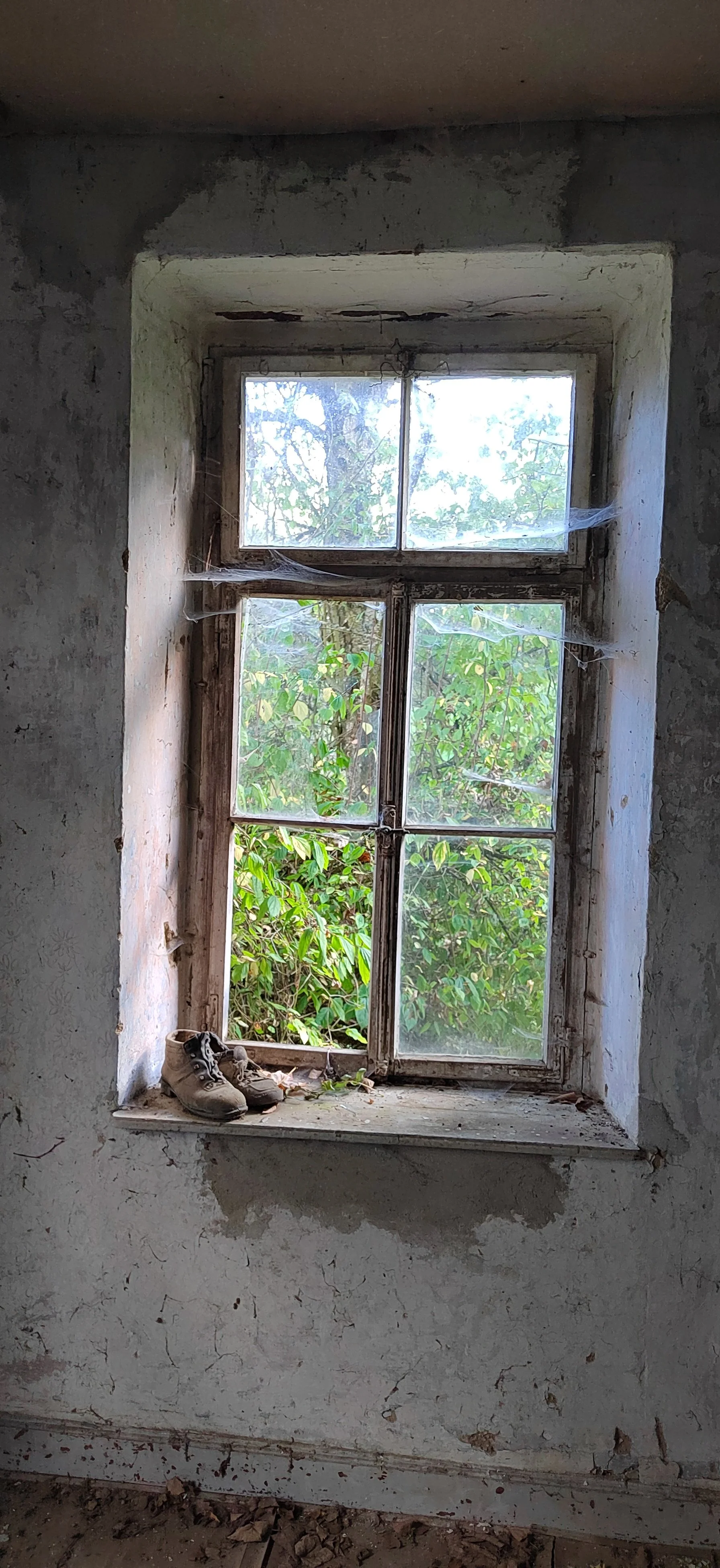 Ein altes, verfallenes Fenster mit vier Scheiben, mit Spinnennetzen und Staub, in einer verlassenen und verfallenen Raum, vor dem Fenster ist ein Paar alter, abgetragener Turnschuhe, im Hintergrund ist grünes Laub zu sehen.