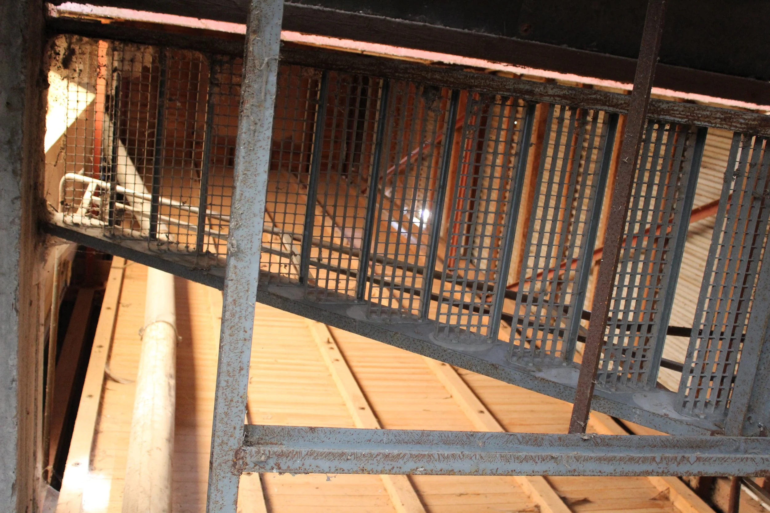 Blick nach oben auf eine Metalltreppe mit Gitterrost in einem Holzhaus, im Hintergrund sichtbare Holzdeckenbalken.