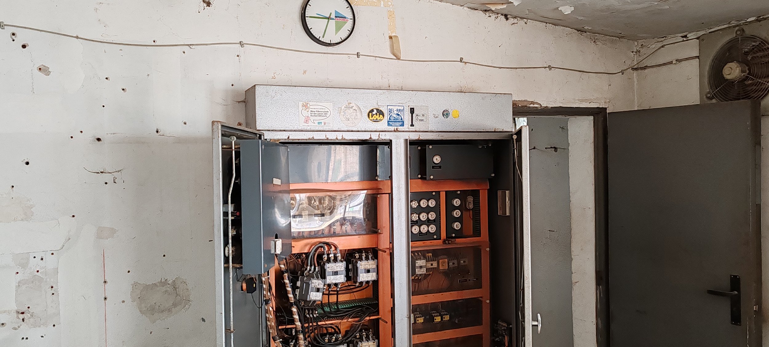 Elektronik- oder Steuerungskasten mit geöffnetem Deckel, zeigt innere Verkabelung und Komponenten, in einem Raum mit beschädigter Wand und einer Uhr an der Wand.
