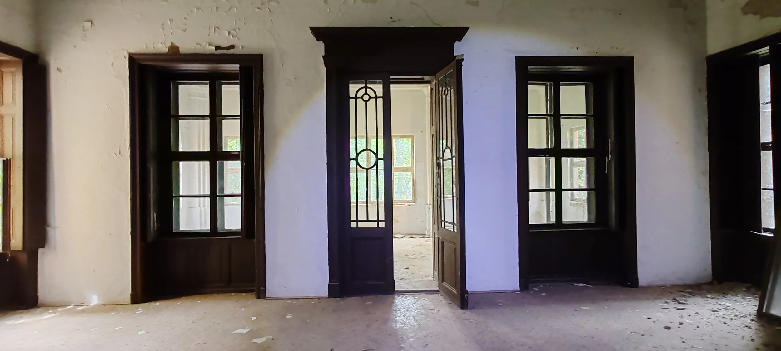 Innenansicht eines verlassenen, alten Hauses mit drei Fenstern und einer geöffneten Tür, die auf einen lichtdurchfluteten Raum hinausführt, Wände sind beschädigt, Staub und Schmutz liegen auf dem Boden.