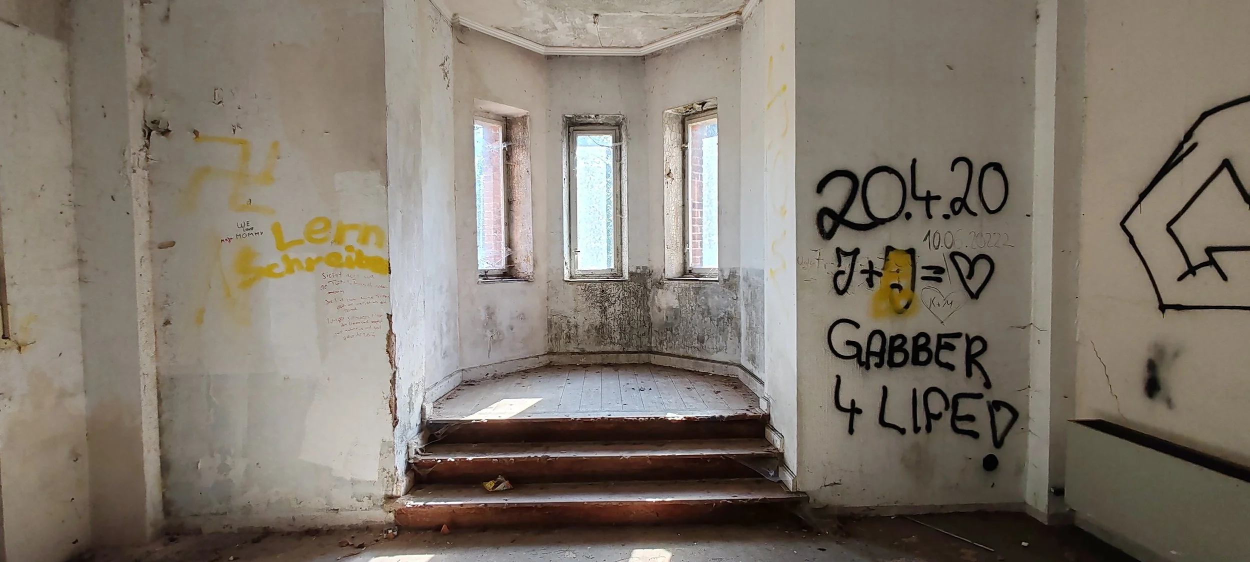 Verlassene, zerfallende Innenraum mit drei Fenstern, grauen Wänden mit Graffiti, Holztreppe, die nach oben führt, und schmutzigem Boden.