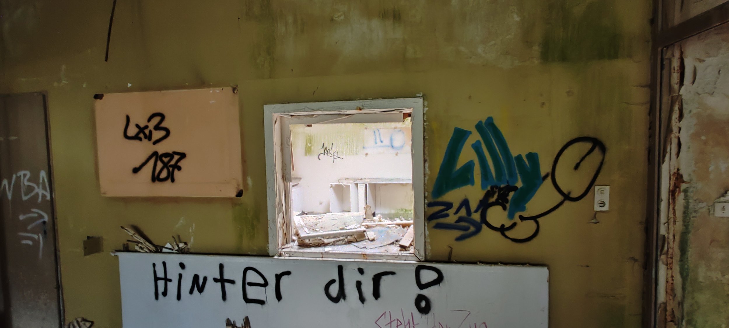 Ein verlassener Raum mit einer kleinen offenen Fensteröffnung, grauer Wand mit Graffiti und Schildern mit deutschen Texten, darunter "Hinter dir!".