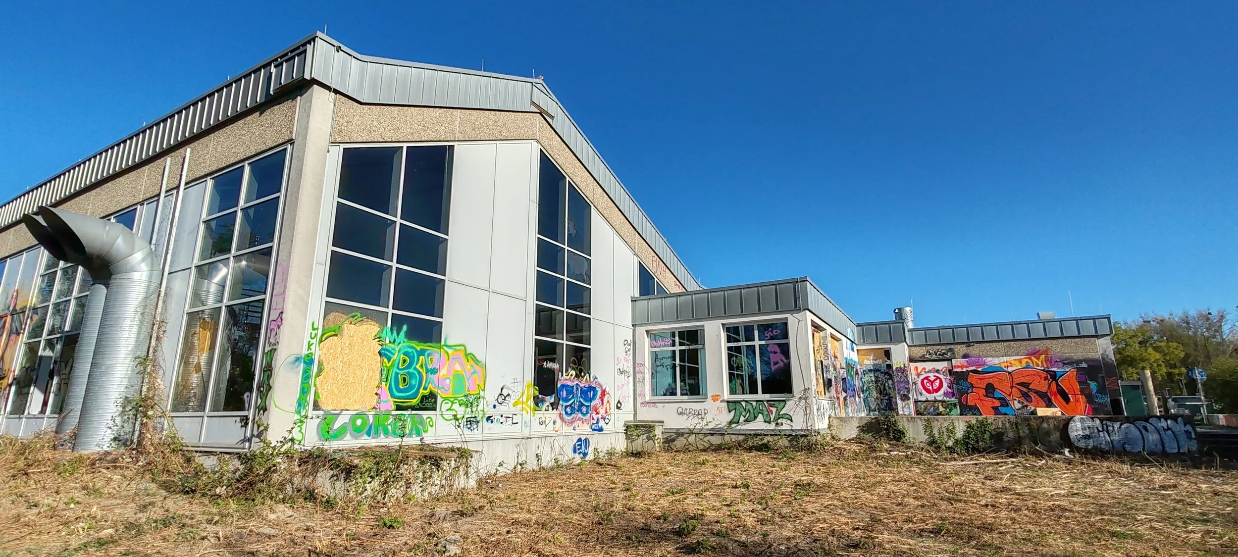 Ein modernes Gebäude mit großen Fenstern, an dessen Wänden graffiti zu sehen sind, auf einer trockenen, unbewachsenen Fläche unter klarem blauen Himmel.