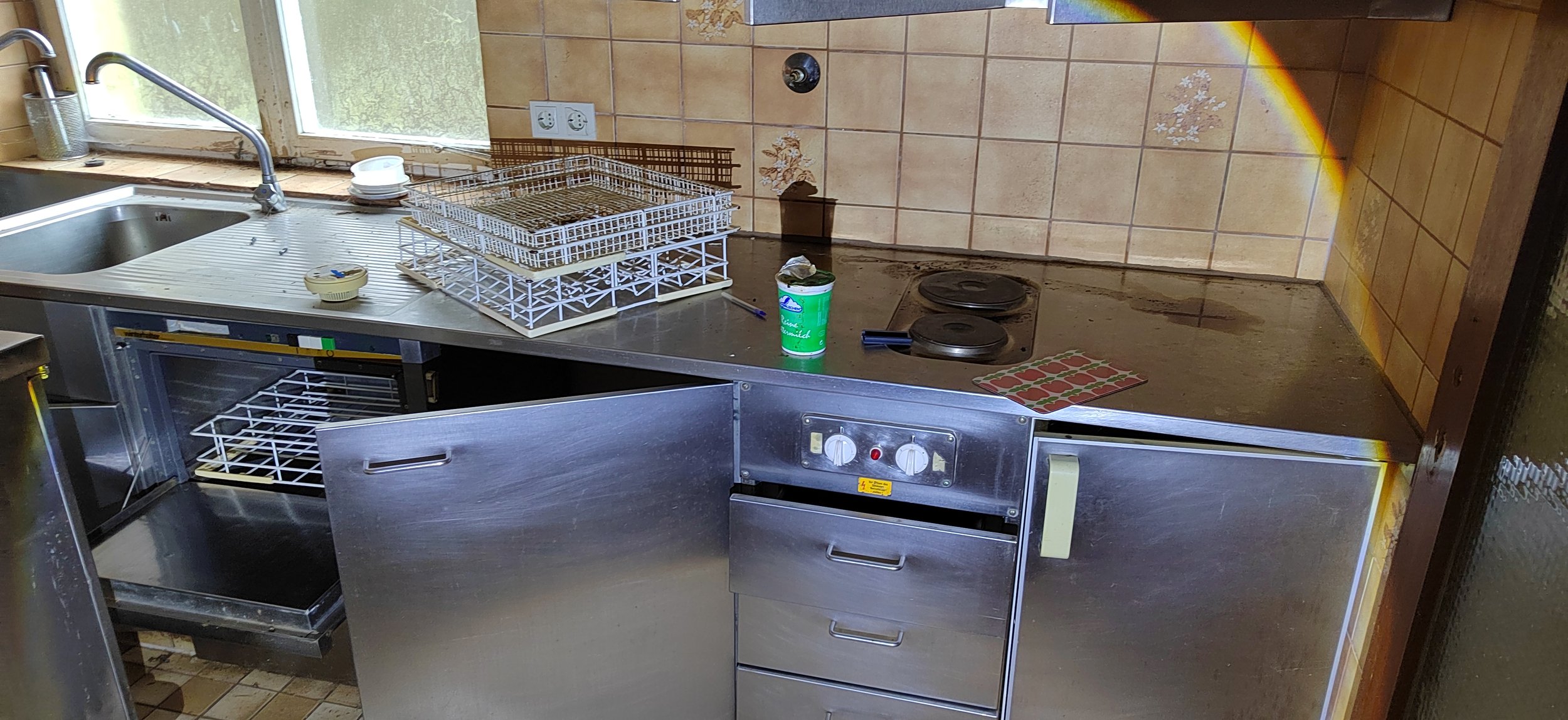 Küchenarbeitsplatte mit Spüle, Geschirrspüler, zwei Kochplatten, und verschiedenen Küchenutensilien, including einem Pott, einem Messer, einem Papiertuch, Geschirr und einem Fenster im Hintergrund.