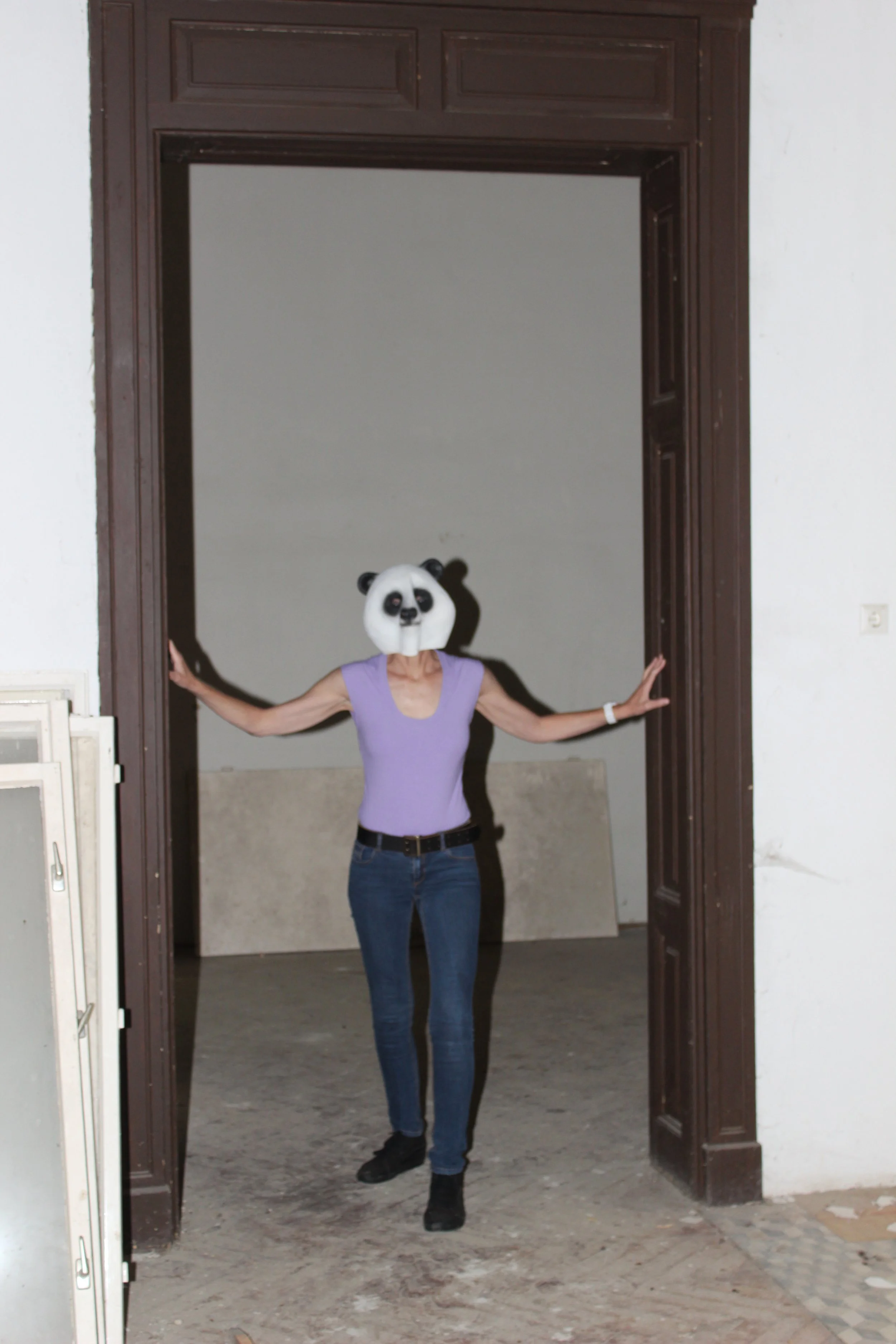 Person in einem Pandamaske, violettem T-Shirt und Jeans steht in einem Türrahmen, die Arme ausgestreckt.