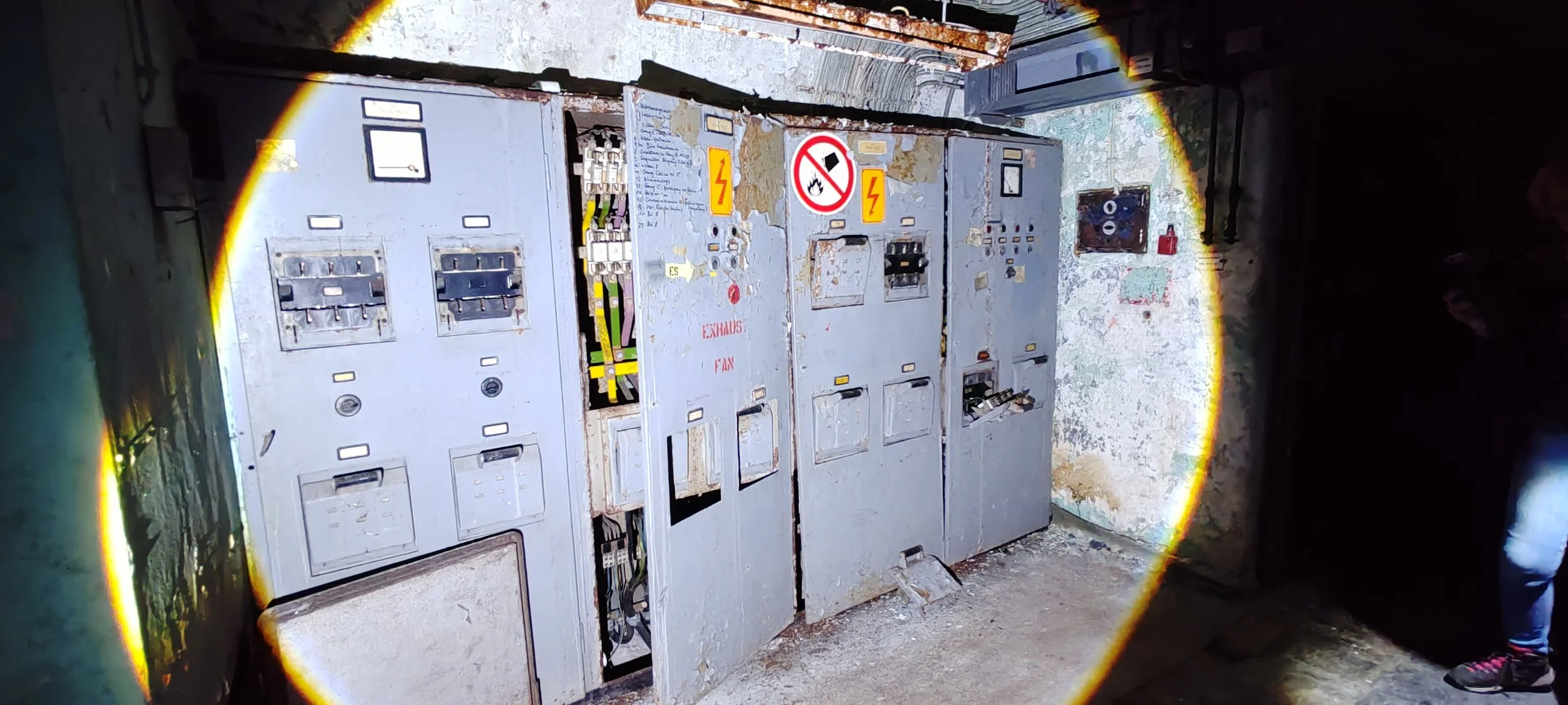 Alter elektrische Schaltkästen in einem dunklen, veralteten Raum, mit Warnhinweisen und geöffneten Teilen, sichtbar durch eine Taschenlampe beleuchtet.