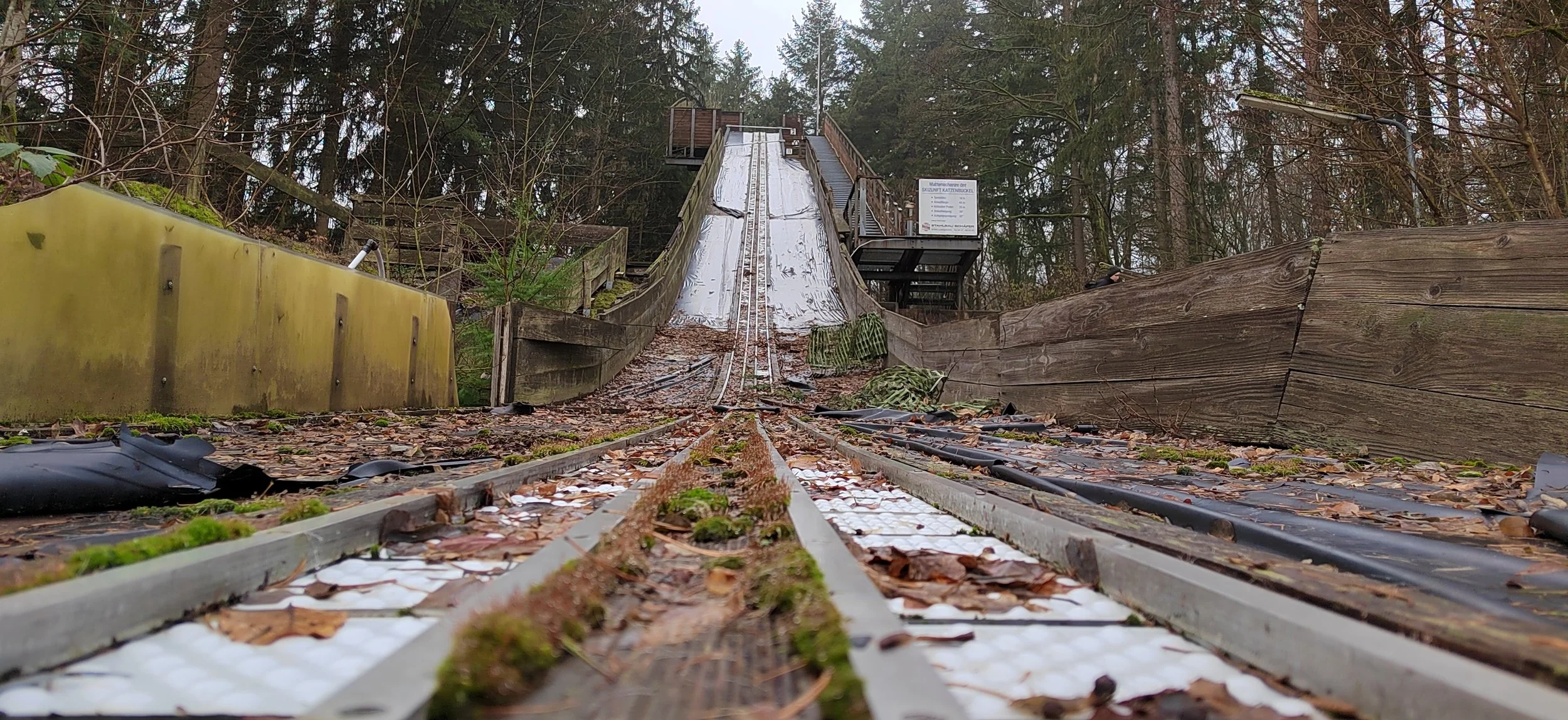 Blick entlang einer Skisprungschanze im Wald, fotografiert aus der Sicht vom Fuß der Schanze, mit Winterschnee und Laub umgeben.