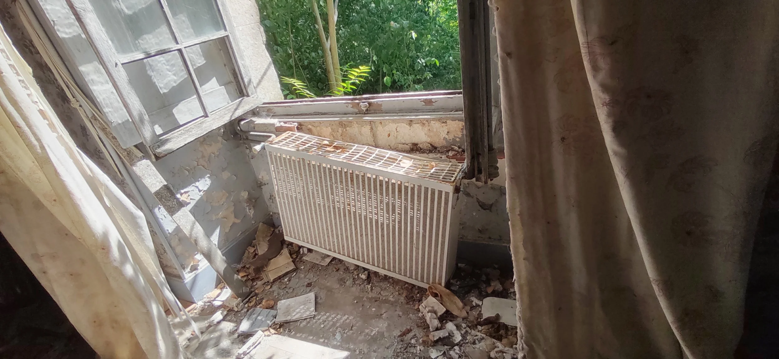 Ein verfallenes, beschädigtes Zimmer mit einem offenen Fenster, einer alten Heizung, zerrissenen Vorhängen und Schmutz am Boden.