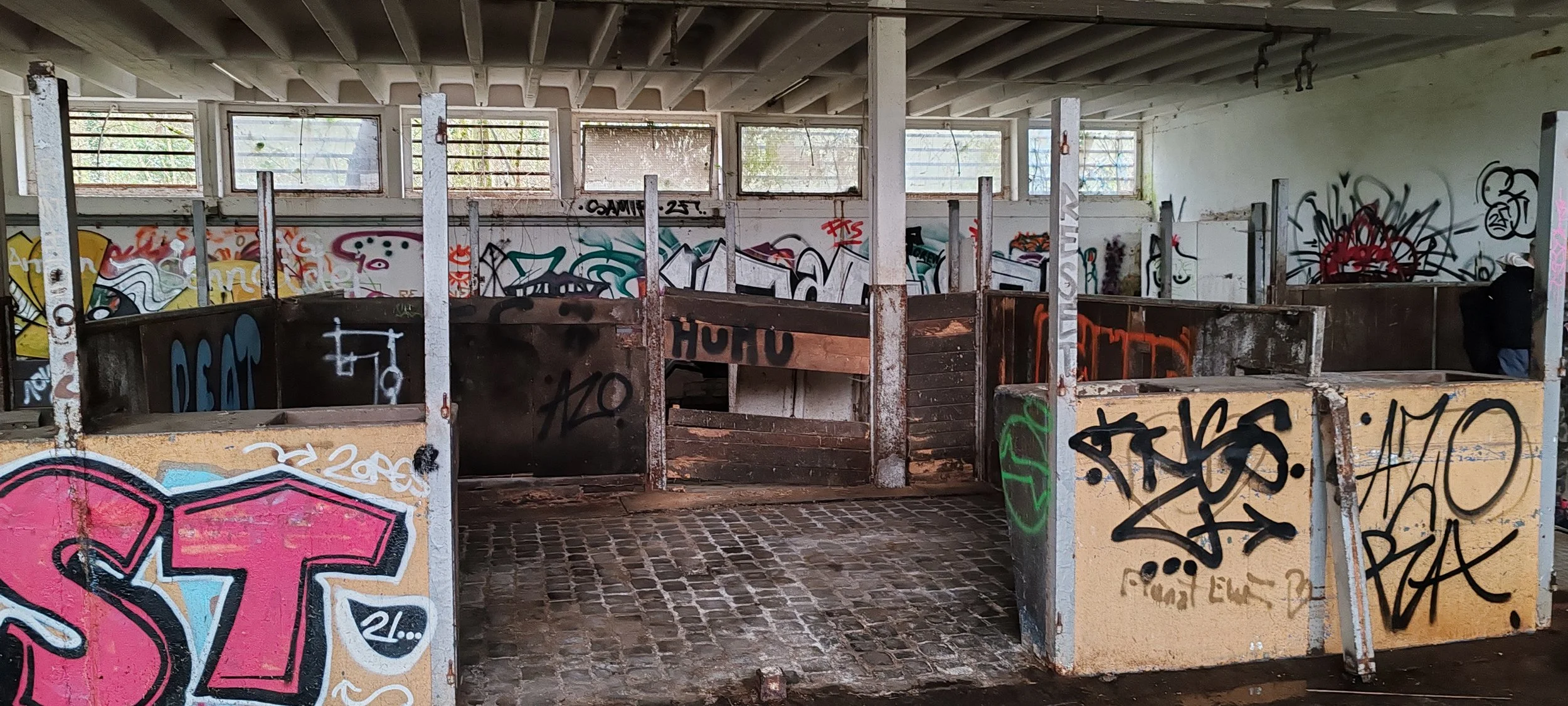 Ein verlassener, graffiti-beschmierter Tennisplatz mit unbenutzten, verwitterten Glaswänden und einer Steinbodenfläche, auf der die Sitzbänke zerfallen sind.