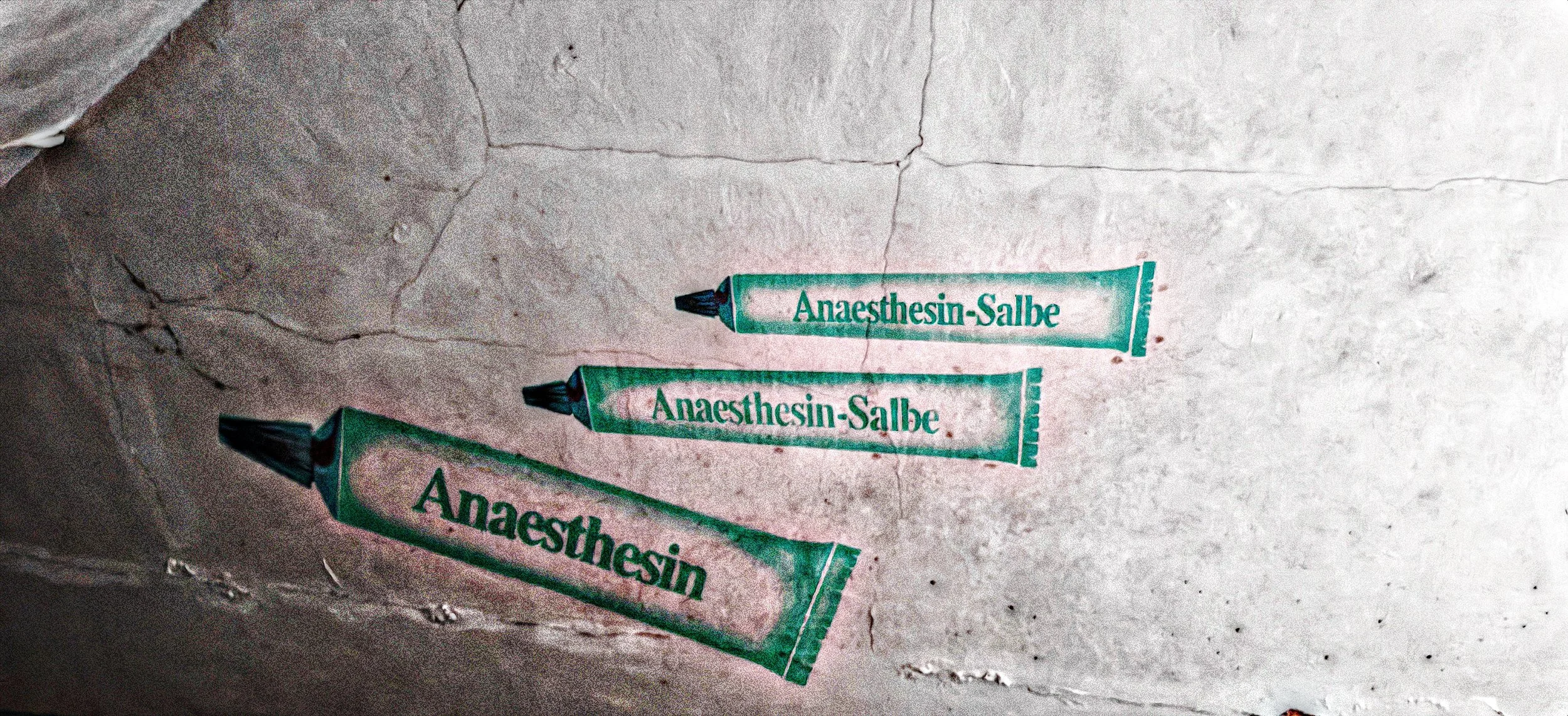 Drei Tuben mit der Aufschrift 'Anaesthesin-Salbe' liegen auf einem grauen Boden.