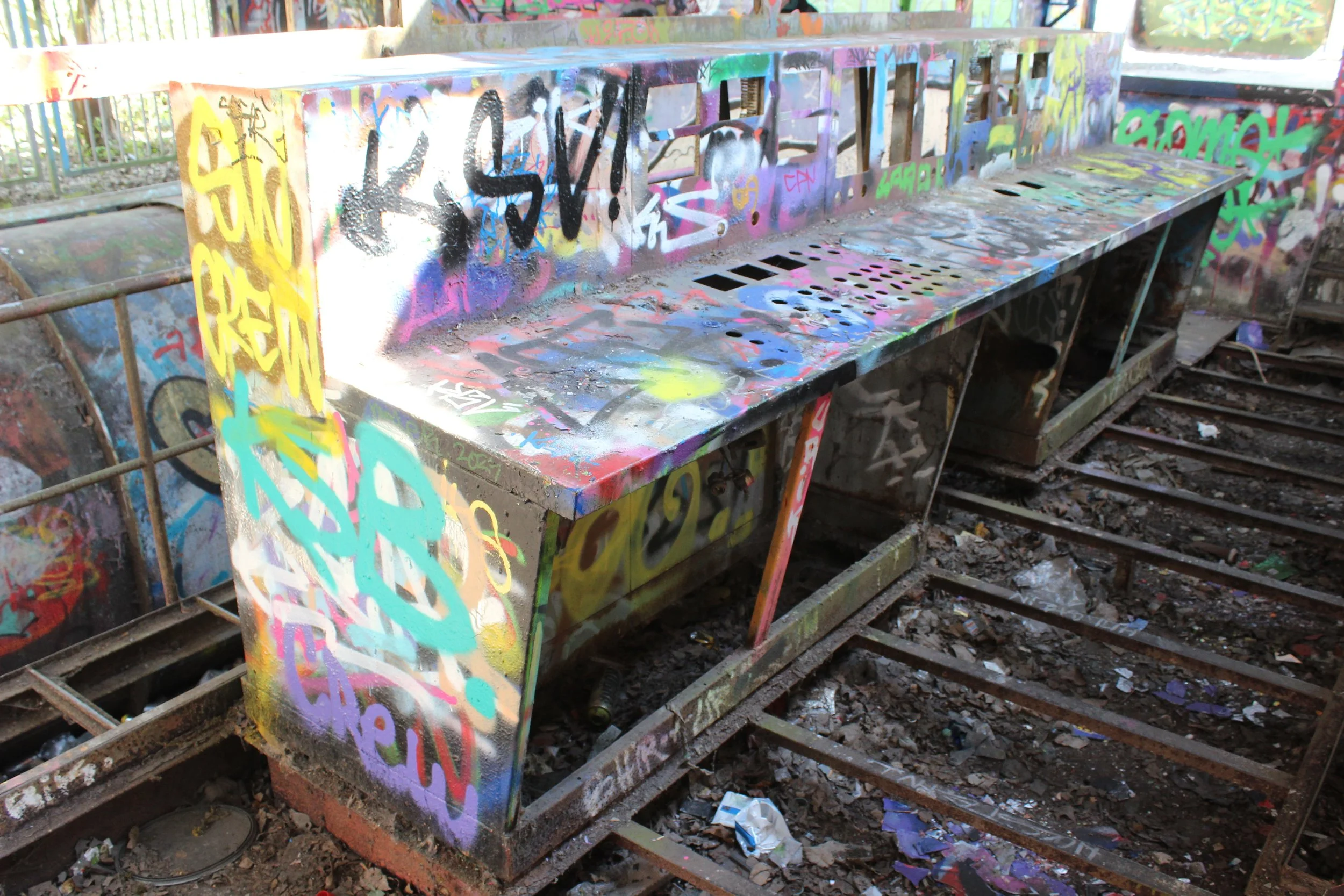 Graffiti bedeckter Dynamobänke auf einem verlassenen, verrosteten Bahnsteig mit Müll auf den Schienen.