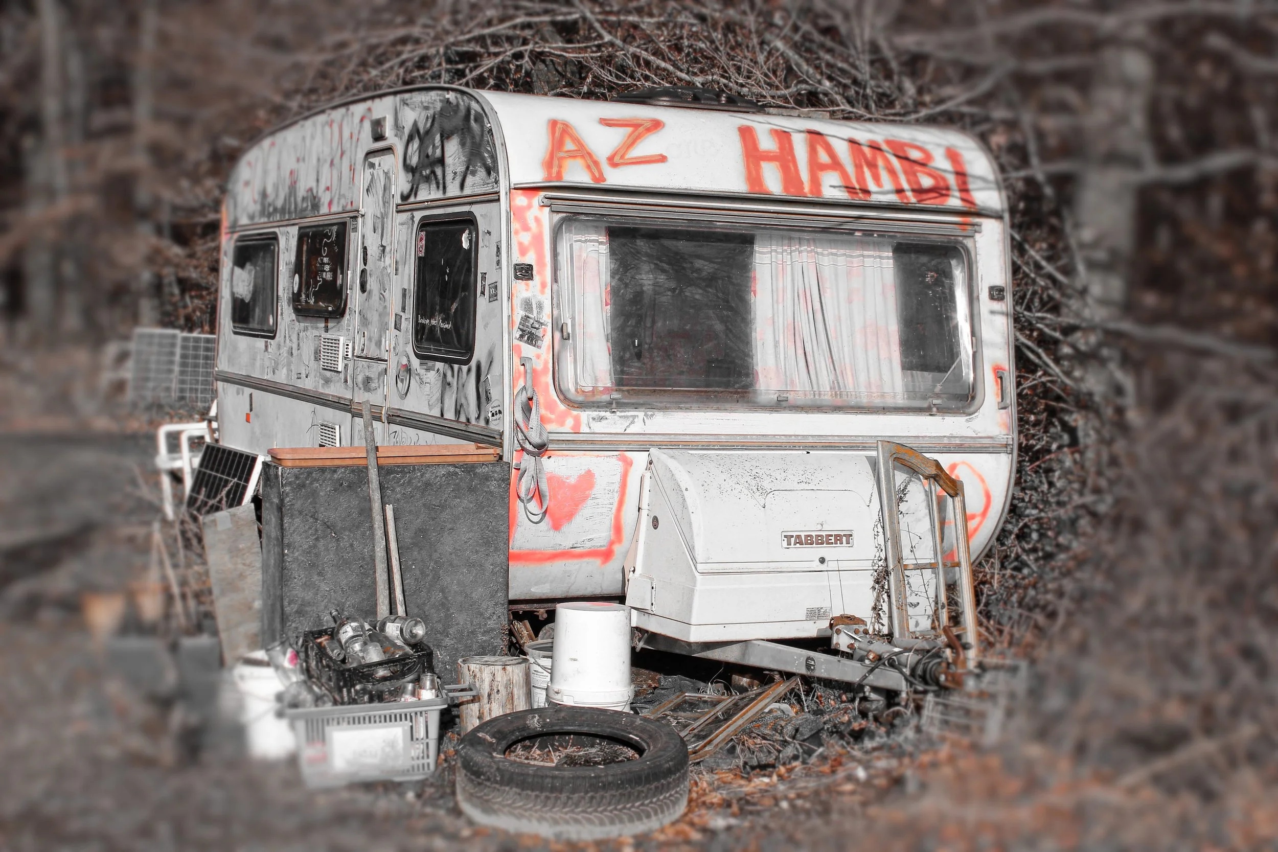 Ein verlassener, graffiti-überschütteter Wohnwagen auf einem unbefestigten, mit Blättern bedecktem Boden, umgeben von alten Gegenständen wie Reifen, Getränkekisten und Werkzeug.