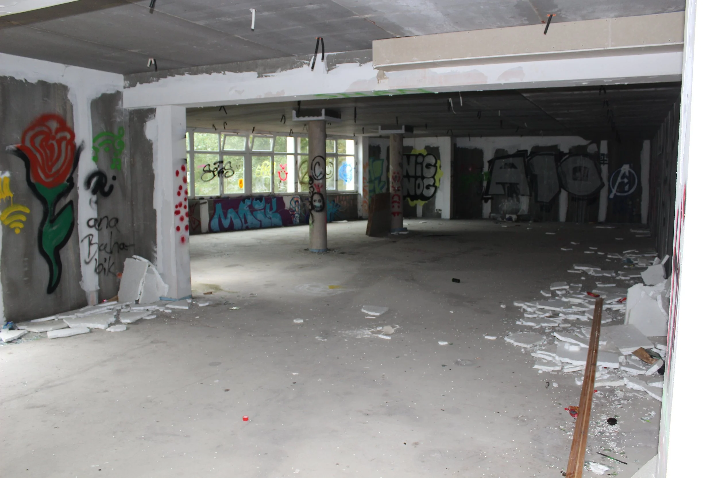 Innenraum eines unfertigen Gebäudes mit Graffiti an den Wänden, großen Fenstern im Hintergrund, Baugerüsten und Müll auf dem Boden.