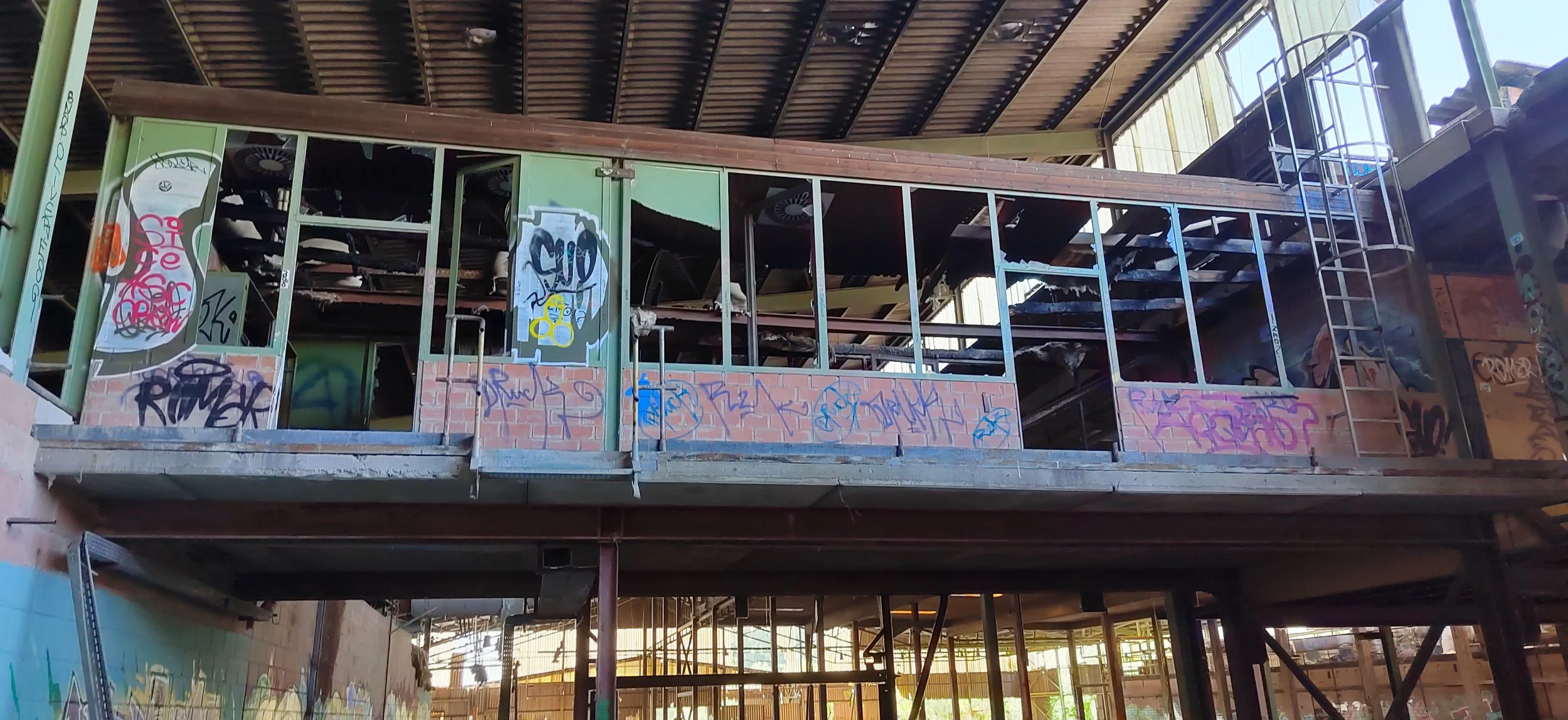 Das Bild zeigt einen zweistöckigen, verlassenen und verunstalteten Gebäudekomplex mit Graffiti an den Wänden. Der obere Stock ist offen und sichtbar durch zerbrochene Fenster und eine Leiter führt zur oberen Ebene.