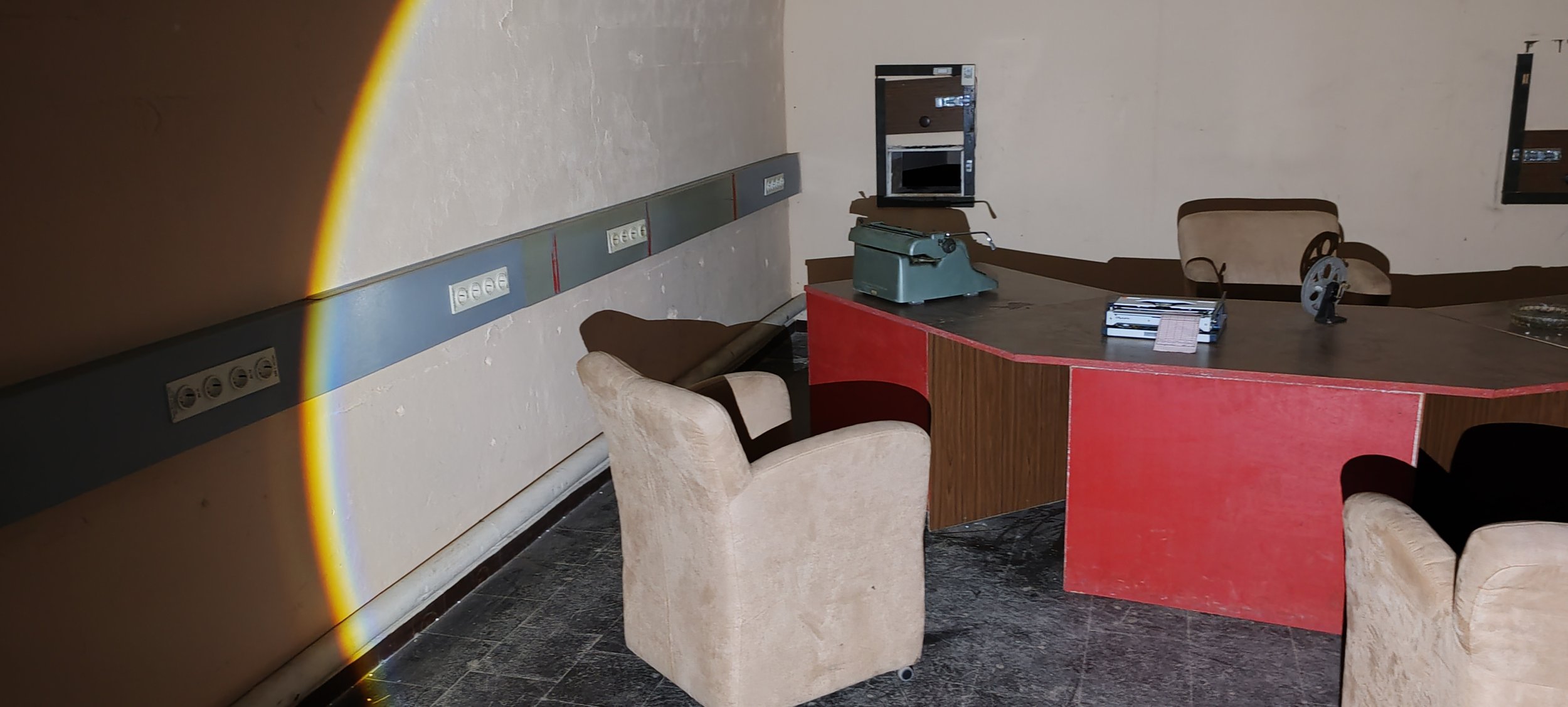 Ein abgelegener Besprechungsraum mit einem großen, halbkreisförmigen Schreibtisch, mehreren gepolsterten Sesseln, einem alten Aktenvernichter, Filmen und Notizen auf dem Tisch, mehreren Steckdosenleisten an der Wand und einem Fenster.