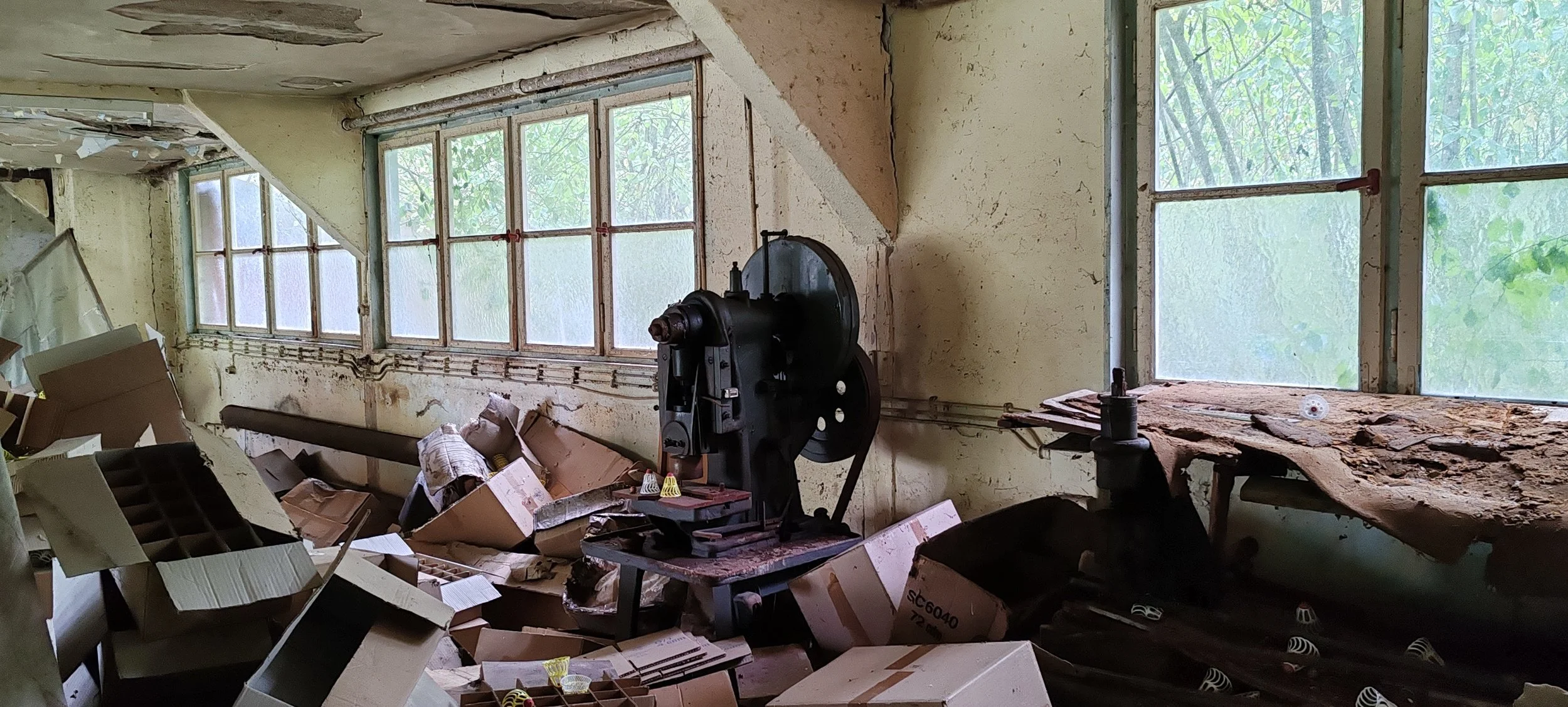Ein verlassener Raum mit unordentlichen Kisten, alten Maschinen und zerbrochenen Fenstern, die auf eine verfallene oder vergessene Umgebung hinweisen.
