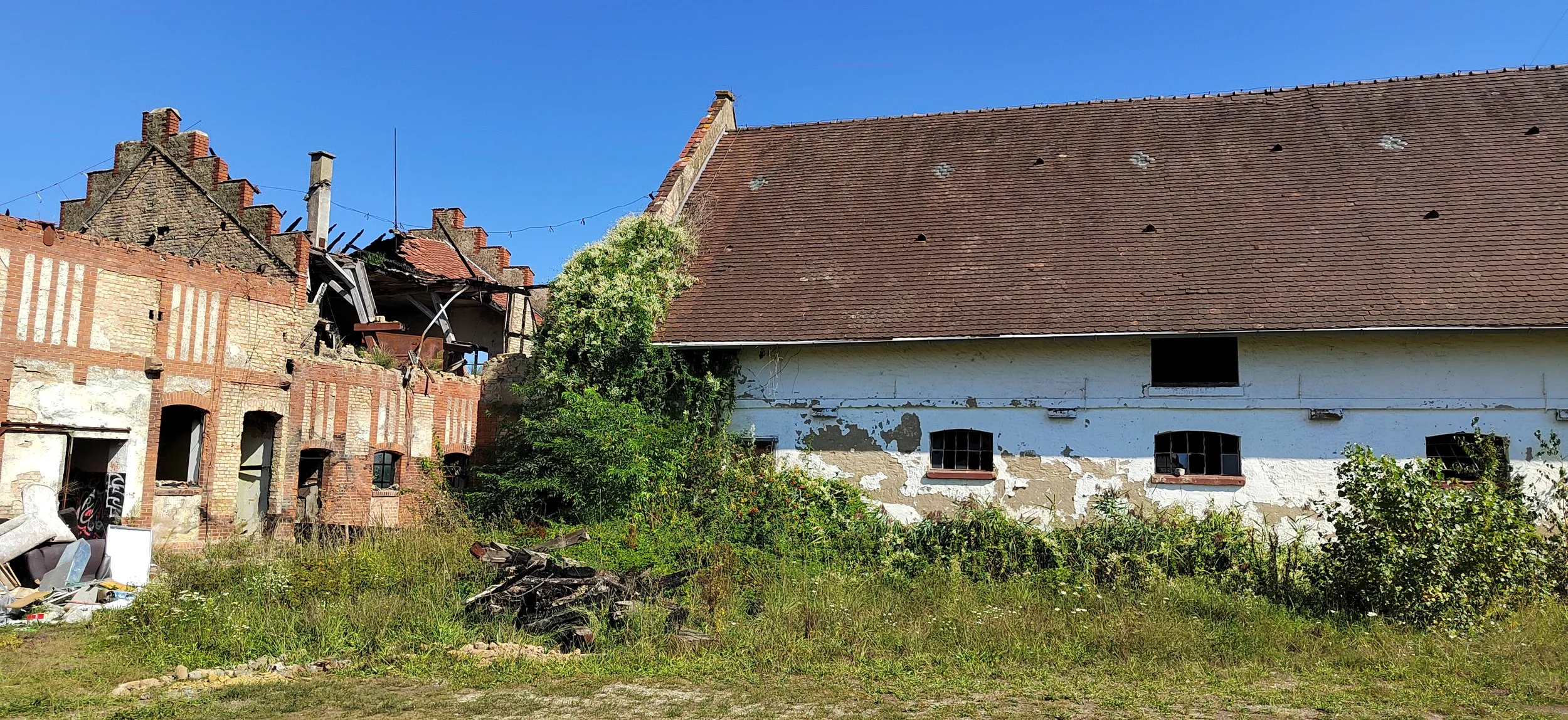 Zwei alte, verfallende Gebäude mit Garten im Vordergrund, eines teilweise eingestürzt, bei sonnigem Himmel.