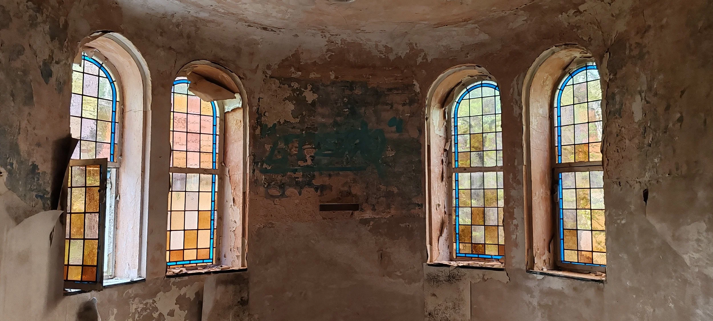 Alte, verfallene Kirchenfenster mit buntem Glas in einer verfallenen Kirche, mit verblasster Wandmalerei im Hintergrund.