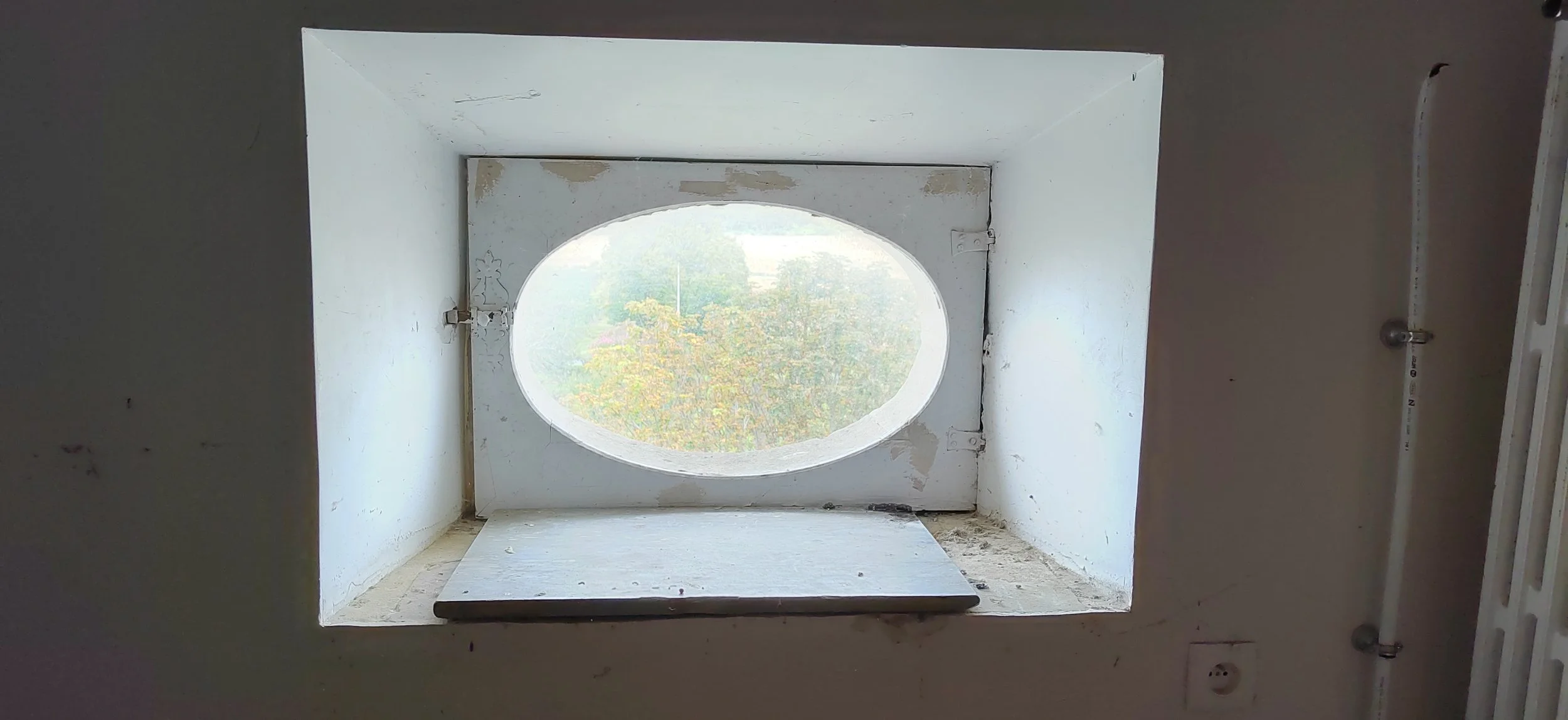 Innenansicht eines kleinen quadratischen Fensters mit ovaler Öffnung, das nach draußen auf eine grüne Landschaft mit Bäumen blickt.