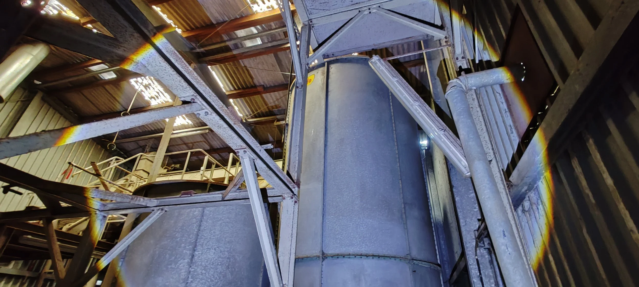 Blick auf eine große industrielle Metallstruktur mit Rohren, Tanks und desigualer Befestigung in einer Lagerhalle.