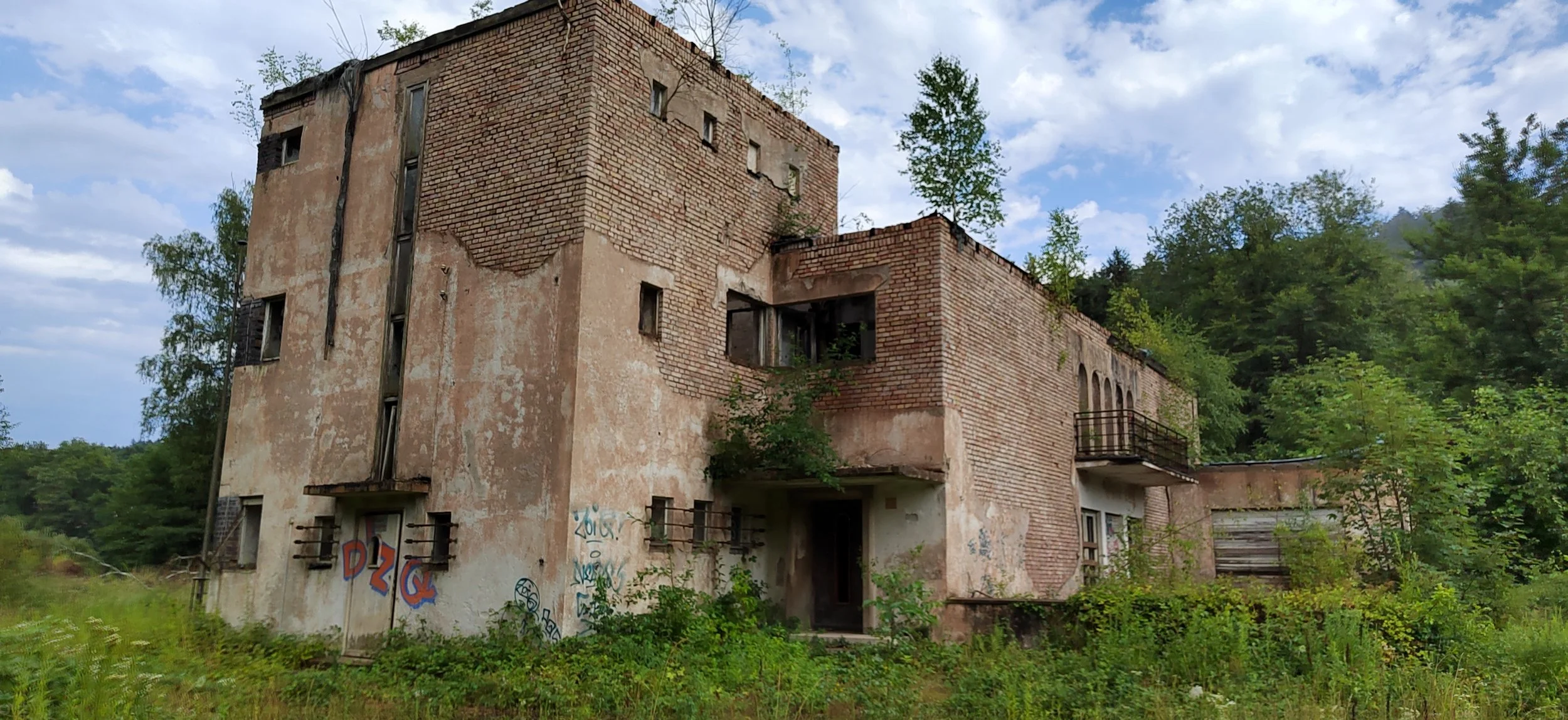 Verfallenes, vernachlässigtes Gebäude mit bröckelnder Fassade, Graffiti an der Wand, umgeben von Grün und Bäumen, bei bewölktem Himmel.