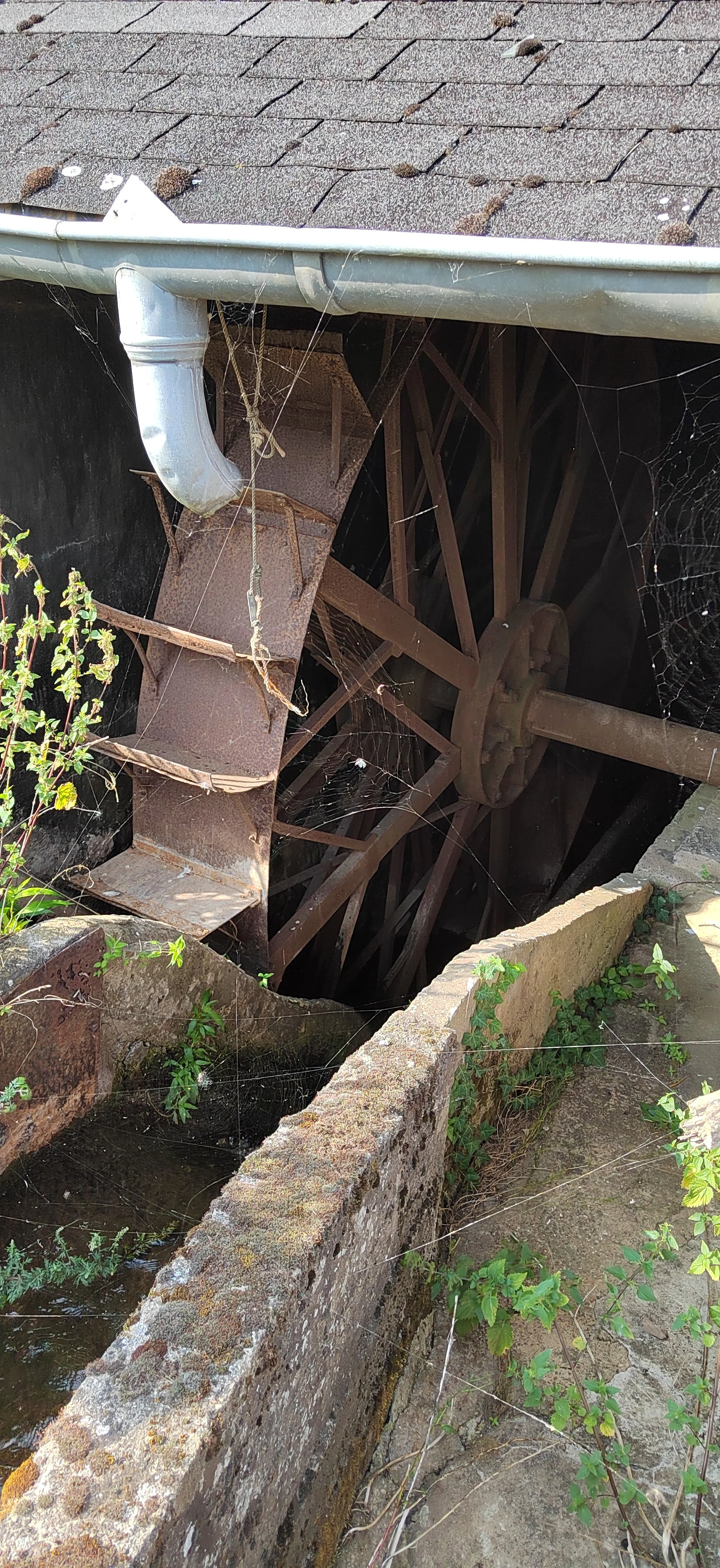 Alter Wasserrad im Garten, von oben fotografiert, mit Spinnweben, rostigen Metallteilen und einem Wasserkanal aus Steinen