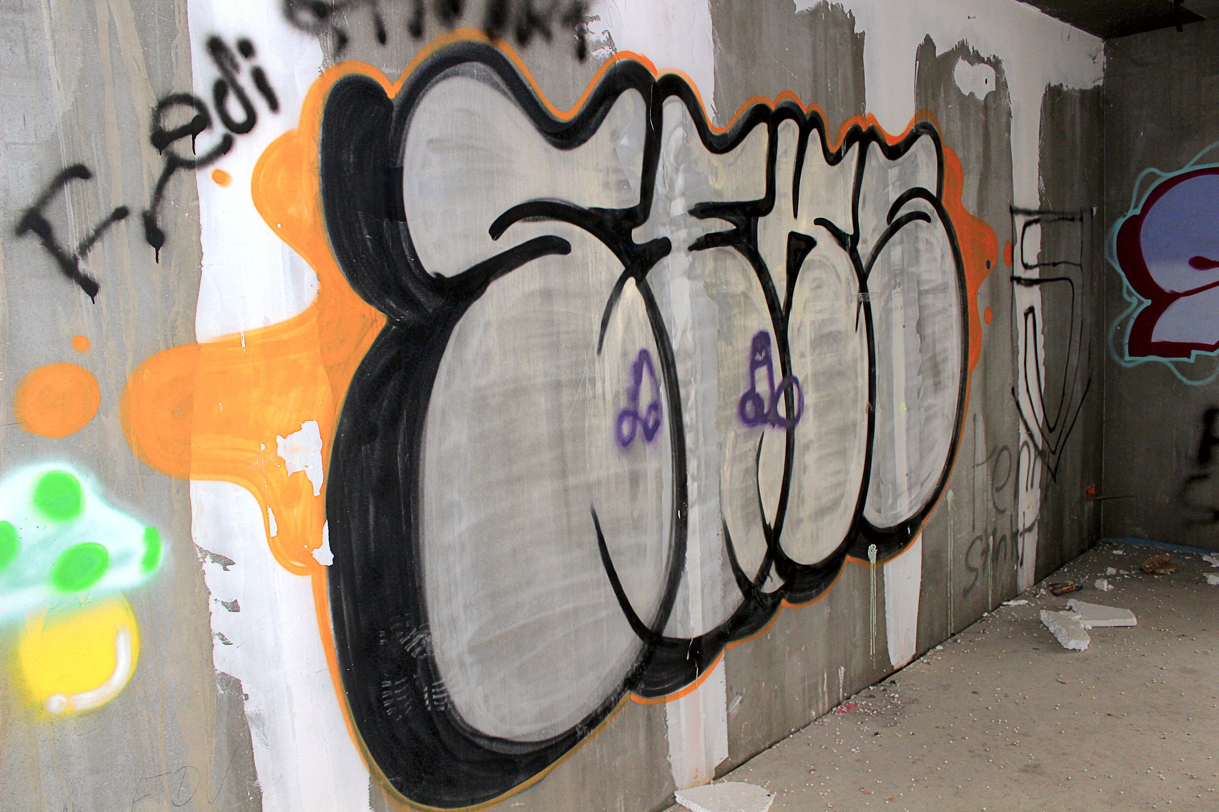 Graffiti auf einer Betonmauer mit schwarzen Konturen, grauen Flächen und orangen Akzenten, sowie einigen kleineren bunten Elementen.