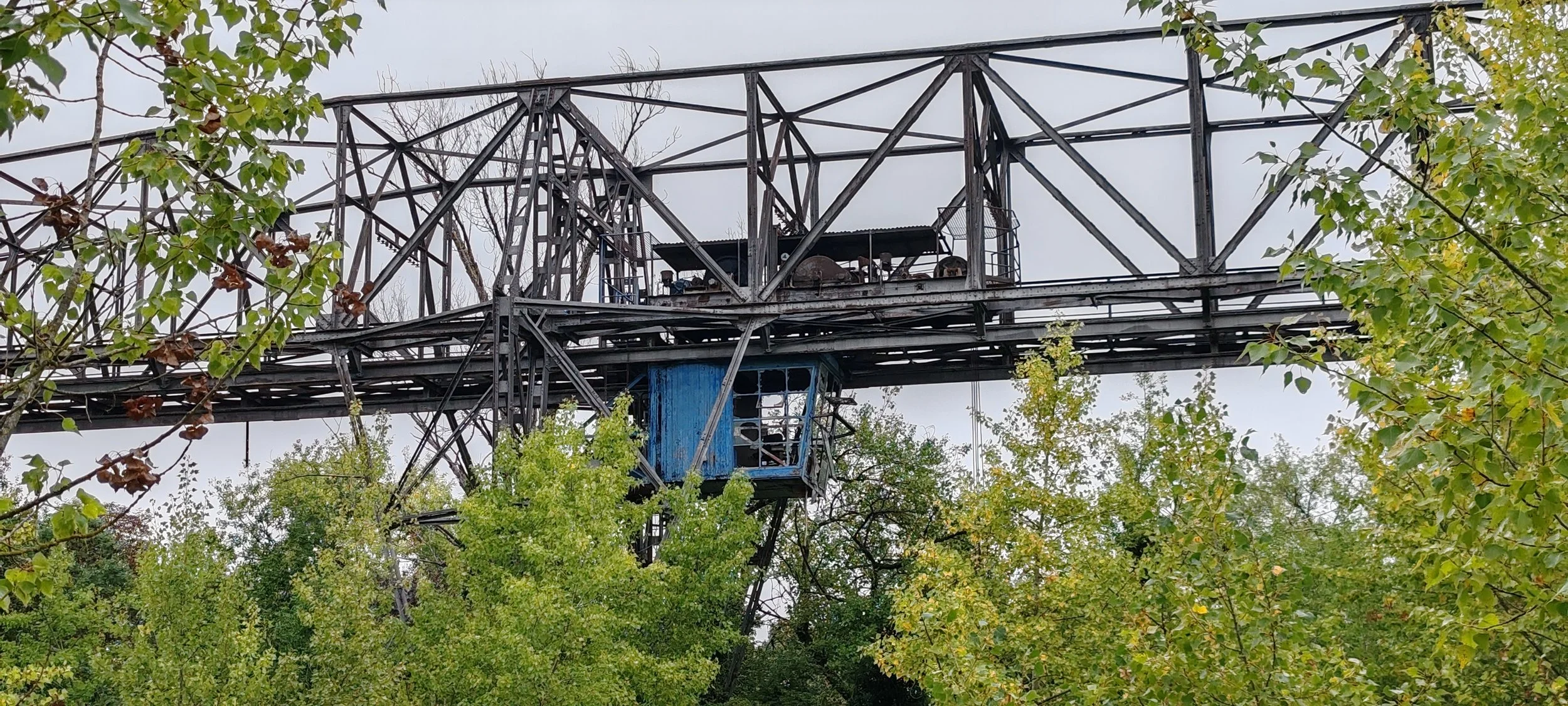 Alter verlassener Industrieschiff mit einer blauen Luke, umgeben von Bäumen, unter einem grauen Himmel.