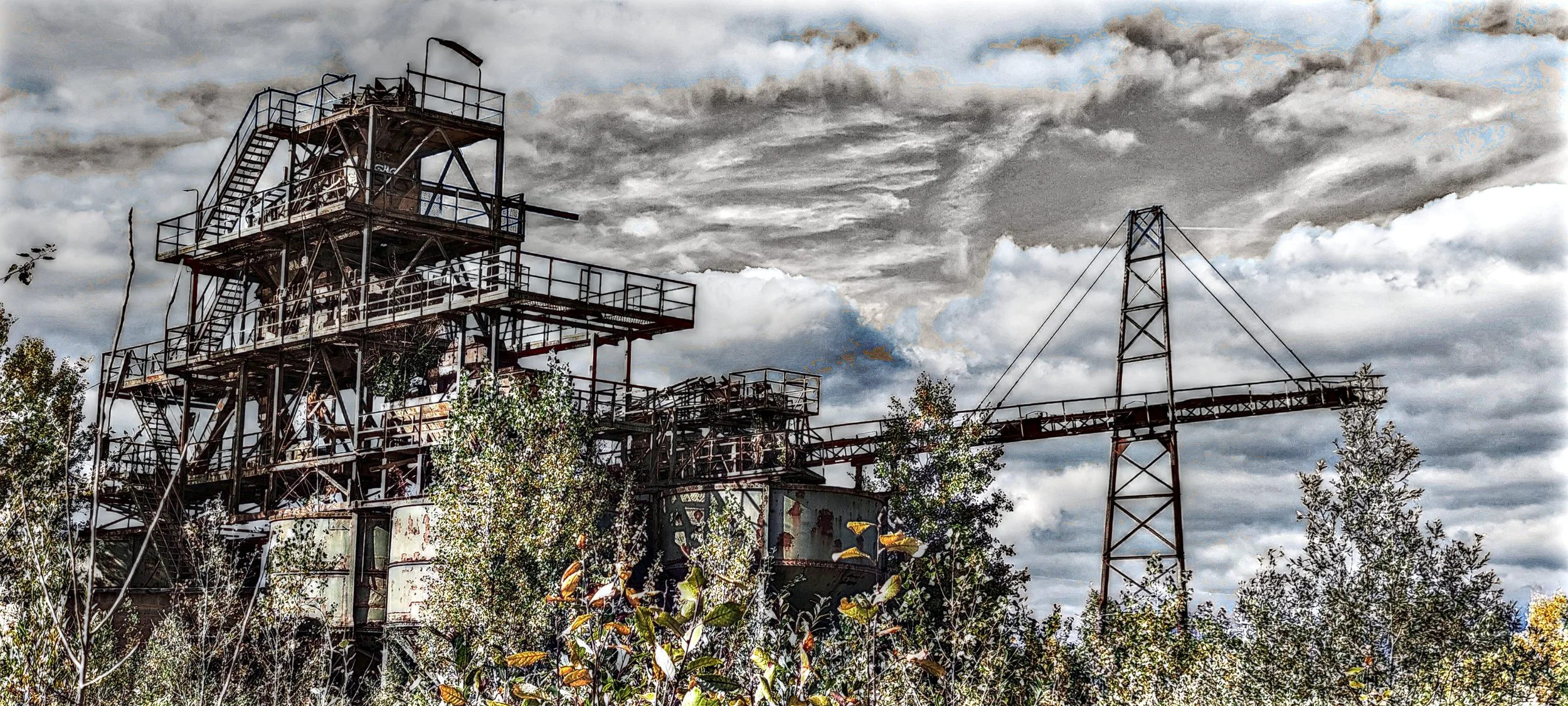 Verlassene, rostige Industrieanlage mit mehreren Ebenen und einem Krans, umgeben von Büschen und Bäumen, unter einem bewölkten Himmel.
