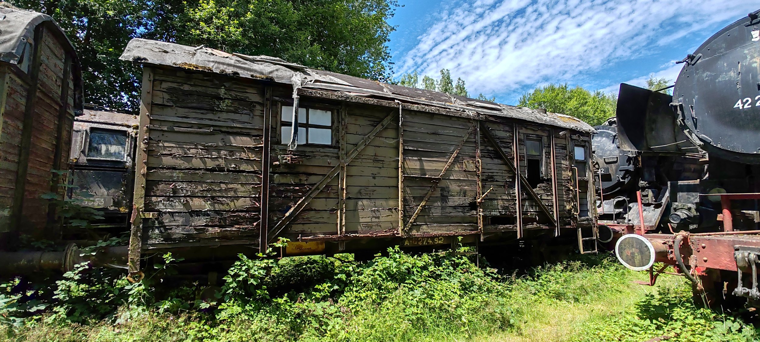 Alter, verrosteter Eisenbahnwagen auf einem Gleis, umgeben von Grün und Bäumen, mit einem blauen Himmel und Wolken im Hintergrund.
