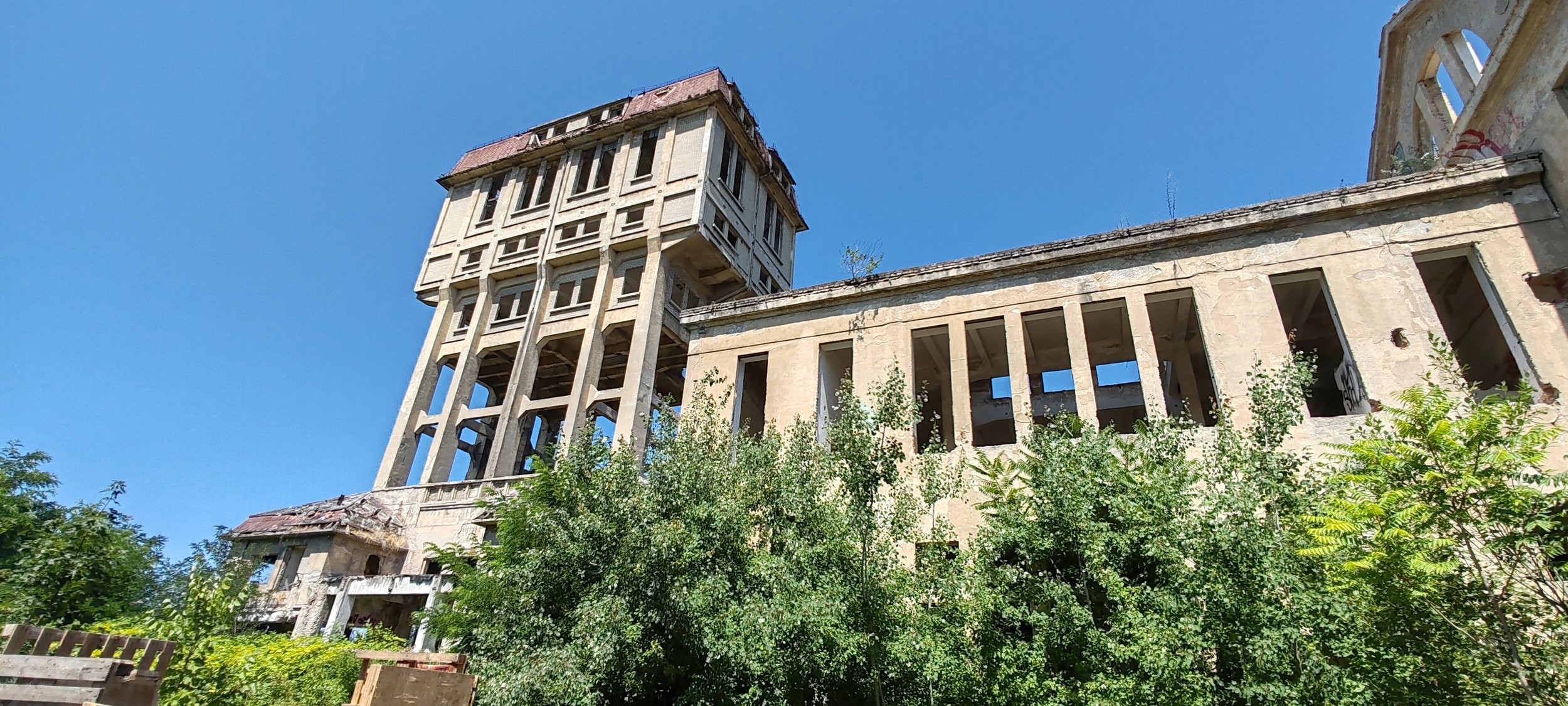 Verfallene und verlassene Gebäude mit einer hohen Wasserleitung oder einem Turm, umgeben von grünen Büschen und einem klaren blauen Himmel.