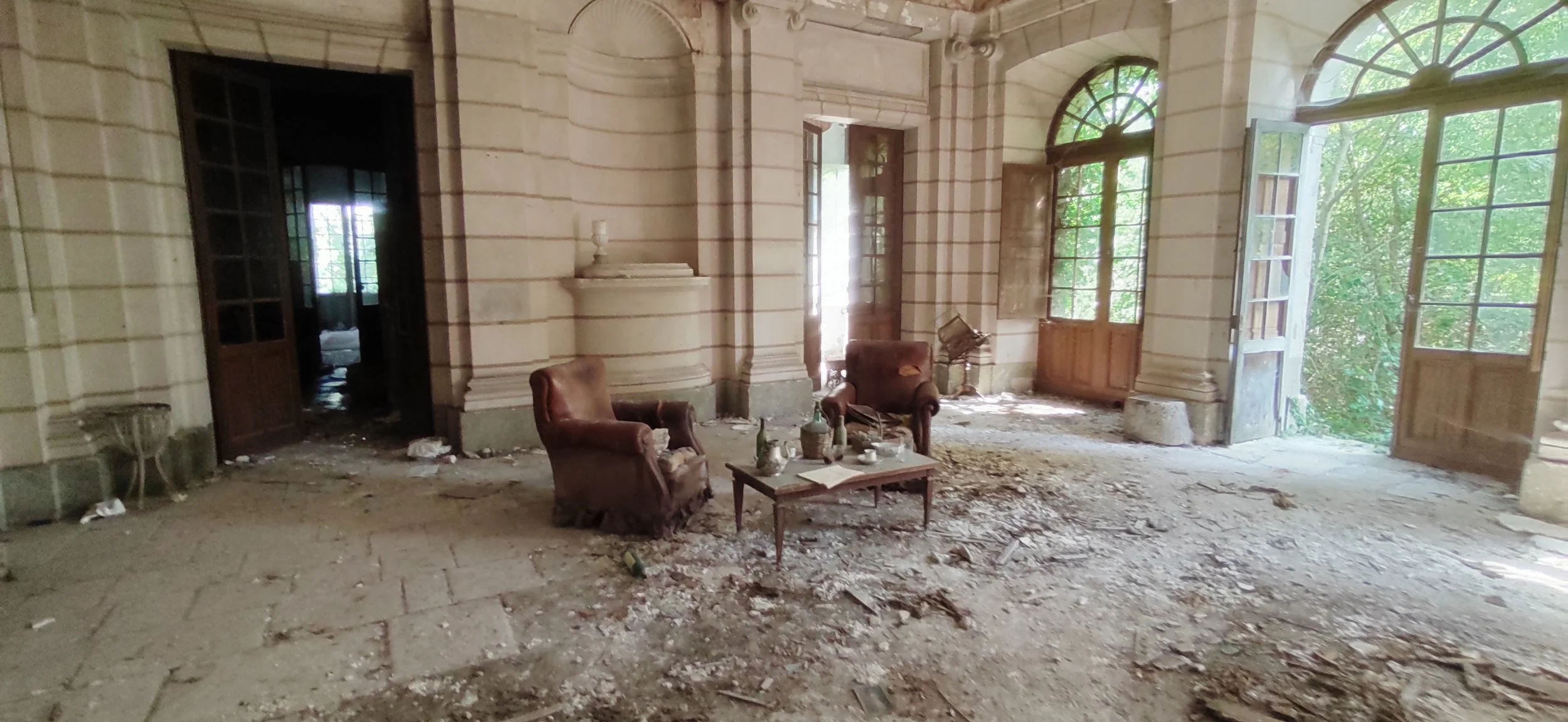 Ein verlassener Raum mit beschädigten Möbeln, Staub, Schutt und offenen Fenstern in einem alten Gebäude.