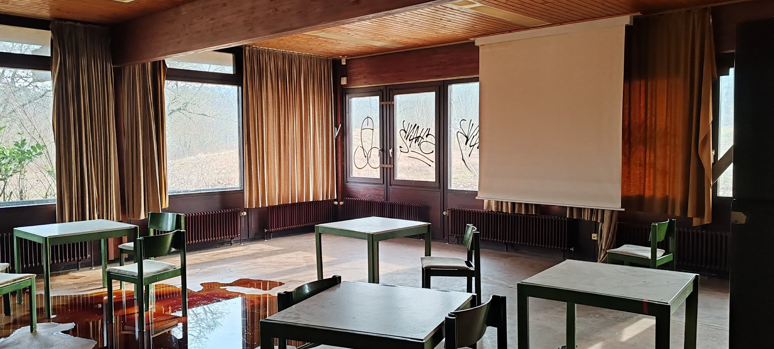 Leeres Zimmer mit Tischen, Stühlen, grauer Leinwand und großen Fenstern mit Goldvorhängen, im Hintergrund sehe ich verschmutzte Scheiben