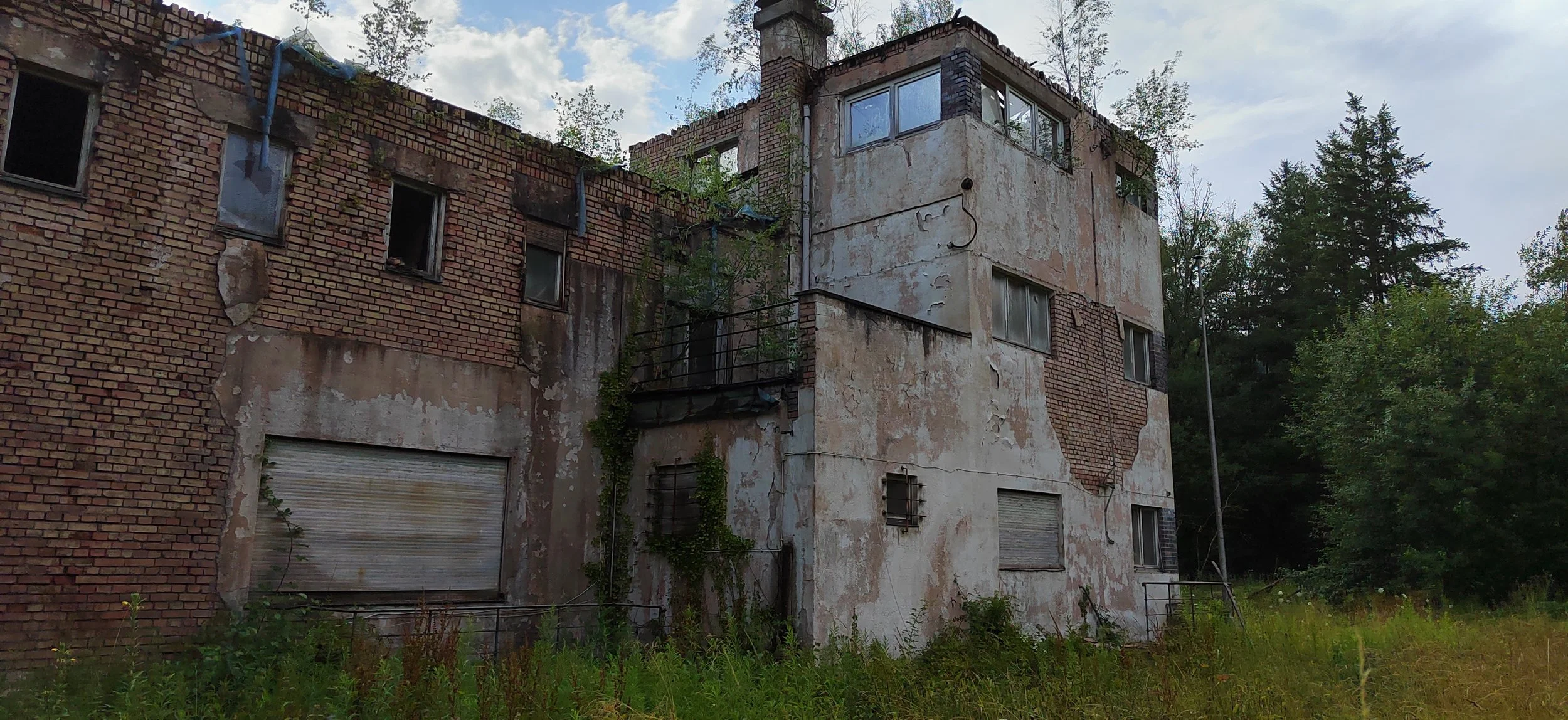 Verfallenes Mehrfamilienhaus mit bröckelnder Fassade, zugewachsenen Fenstern und eingezäunten, heruntergekommenen Balkonen in einer bewachsenen Umgebung mit Bäumen und Wiesen.