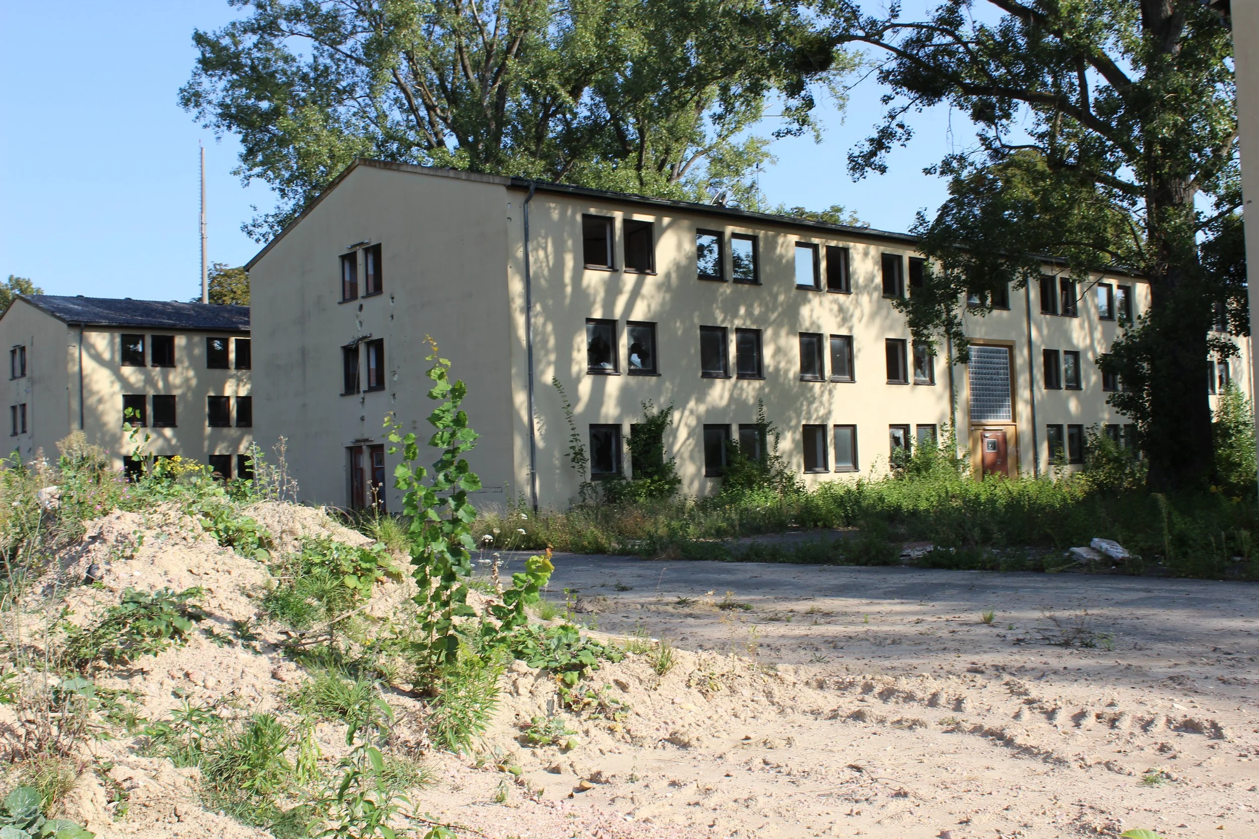 Ein altes, mehrstöckiges Mehrfamilienhaus mit mehreren Fenstern, umgeben von Bäumen und unbefestigtem Boden.