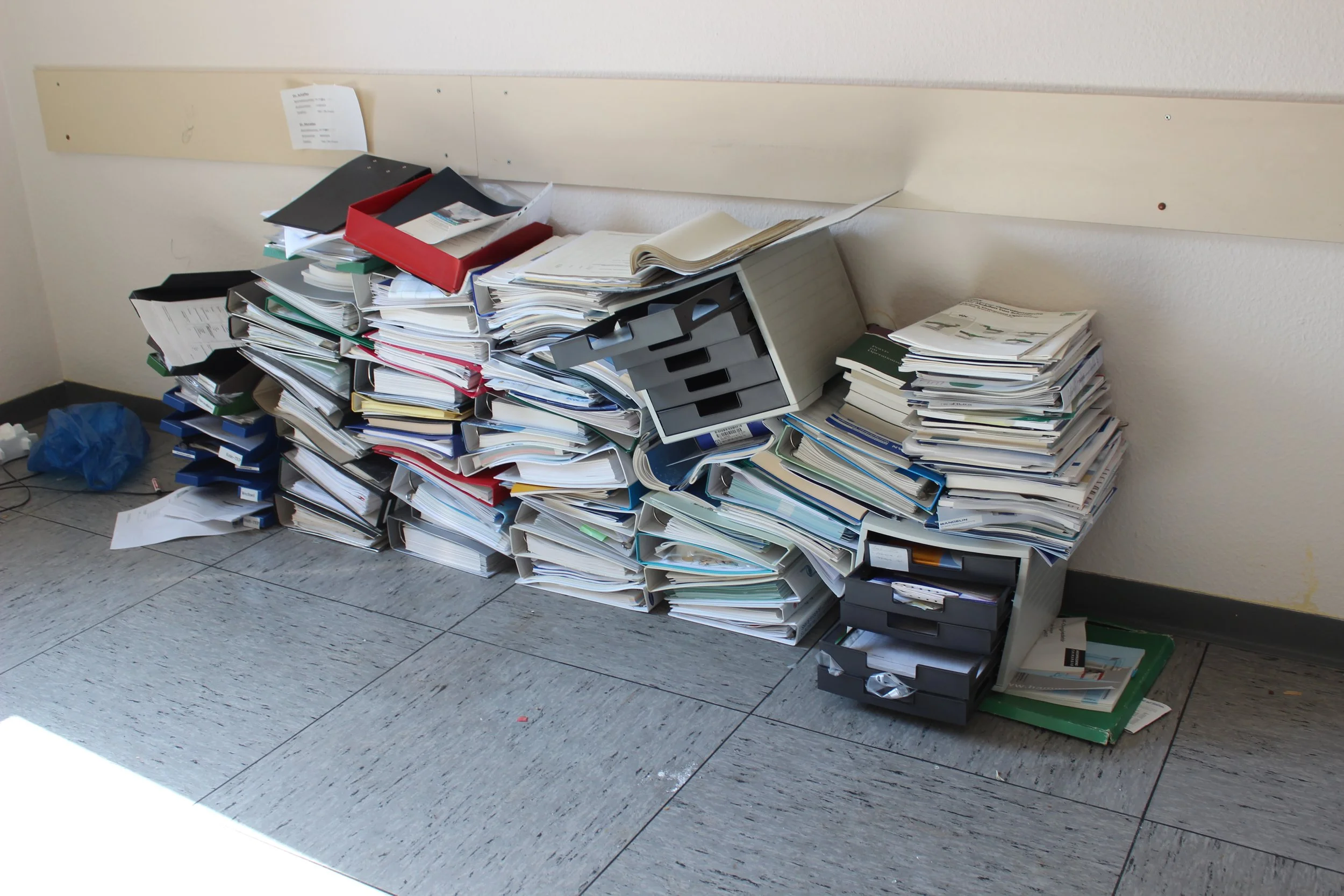 Unordentliche Büroablage mit vielen Aktenordnern, Papieren und Papierstapeln auf dem Boden in einem Büro.
