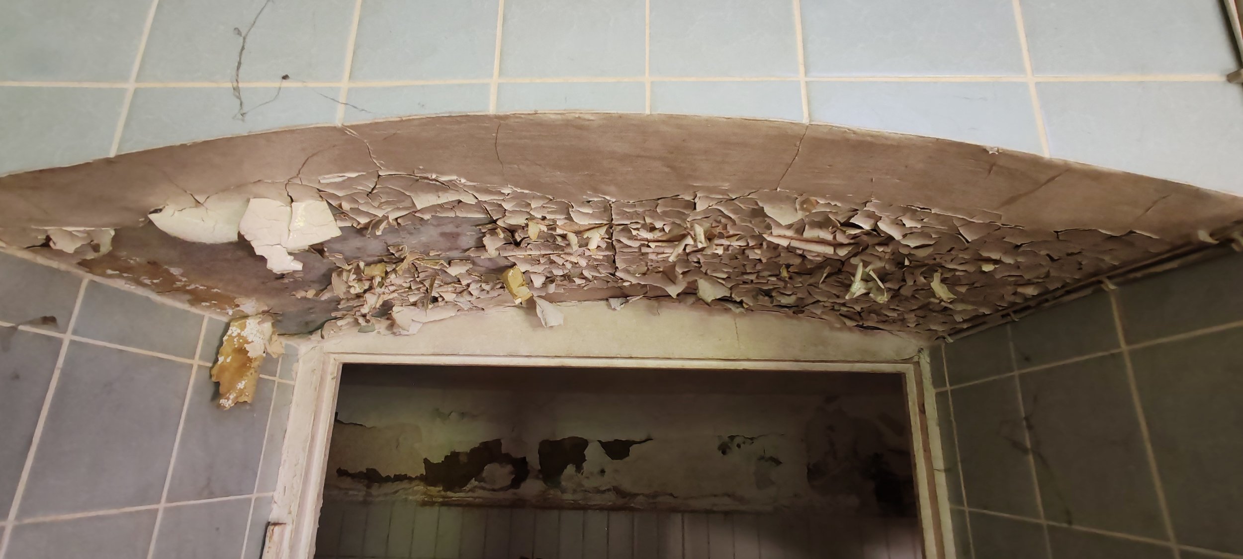Decke mit abblätternder, alter Putz und Schäden in einem Raum mit Fliesenspiegel.
