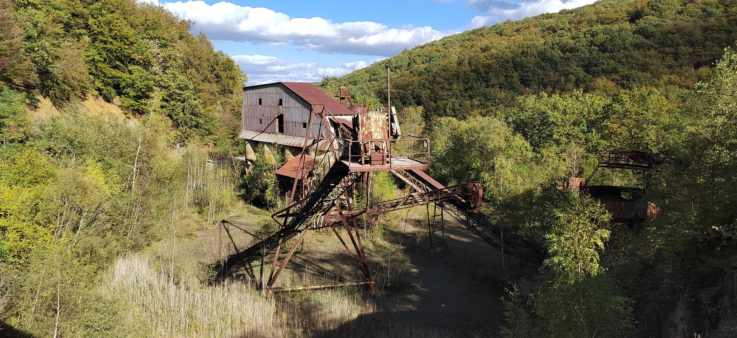 Verrostete verlassene Bergbauanlage in einer grünen, bewaldeten Hügellandschaft, unter blauem Himmel mit Wolken.